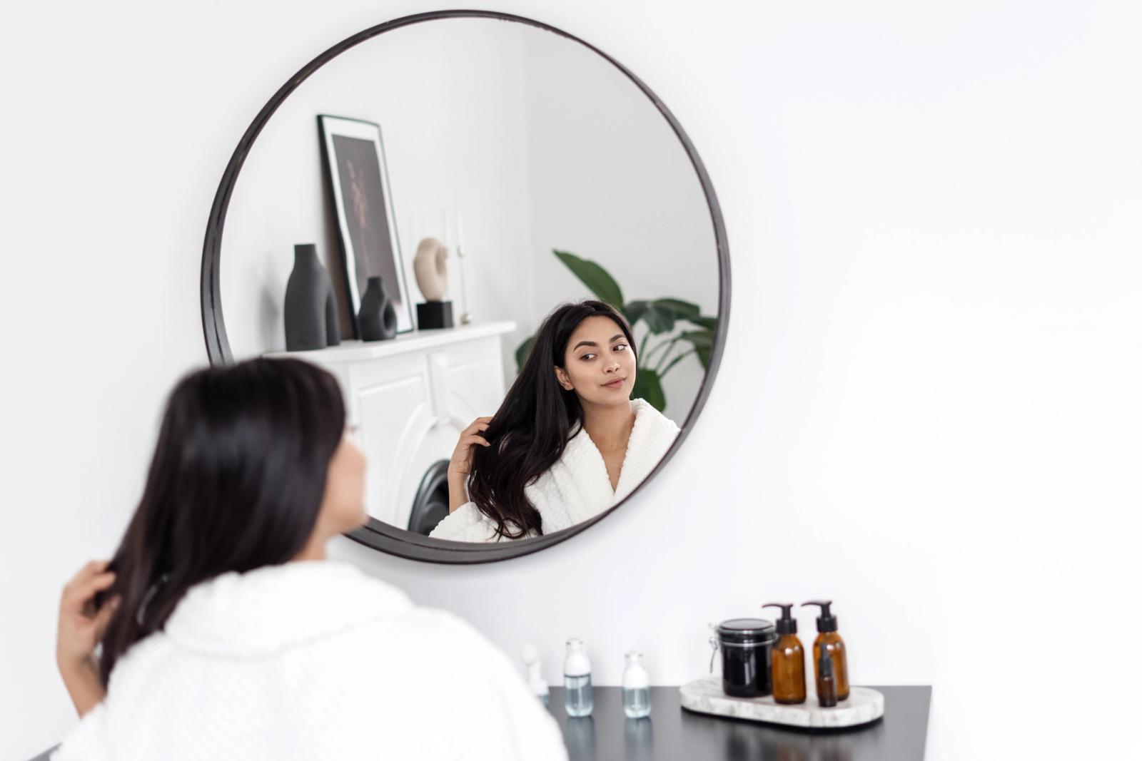 Woman sits at a vanity, looking at her reflection whilst she smiles and combs hair with her fingers