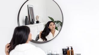 Woman sits at a vanity, looking at her reflection whilst she smiles and combs hair with her fingers