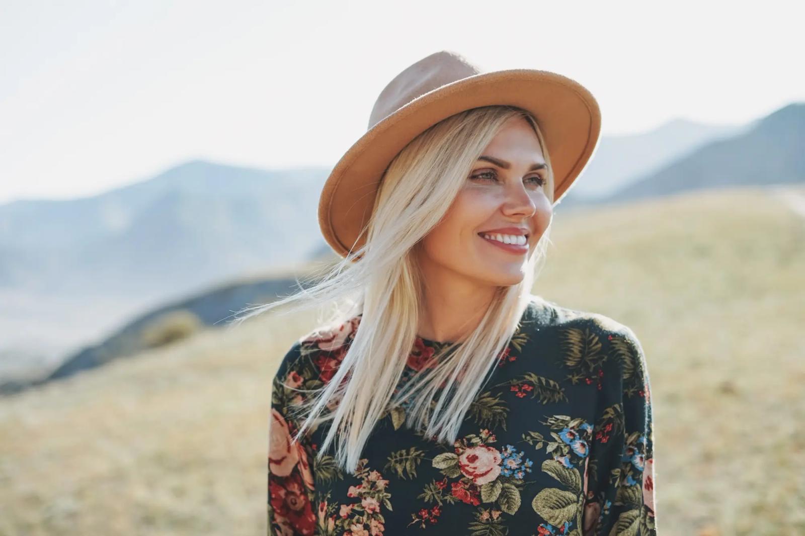 Woman with blonde hair in a beige hot standing in a field in the mountains smiling and looking out to the views.