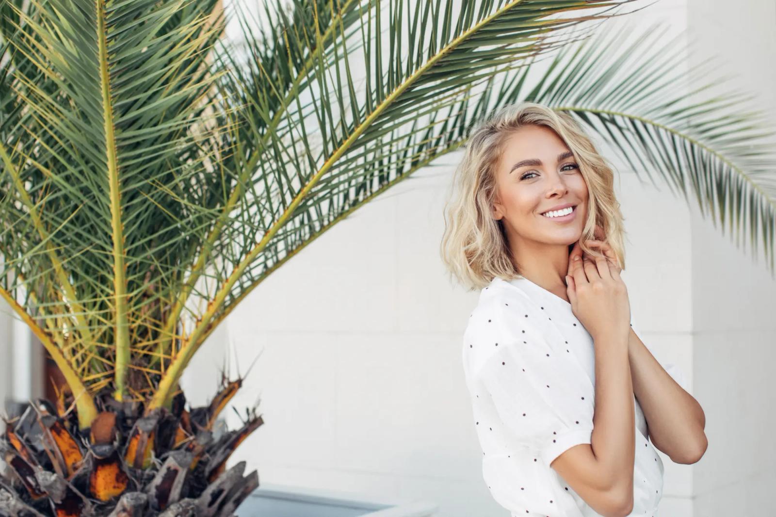 Blonde woman with wavy short hair stands on her left side standing at the camera next to a palm tree