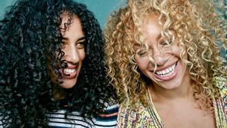 Woman with curly black hair and woman with blonde curly hair laugh standing beside each other