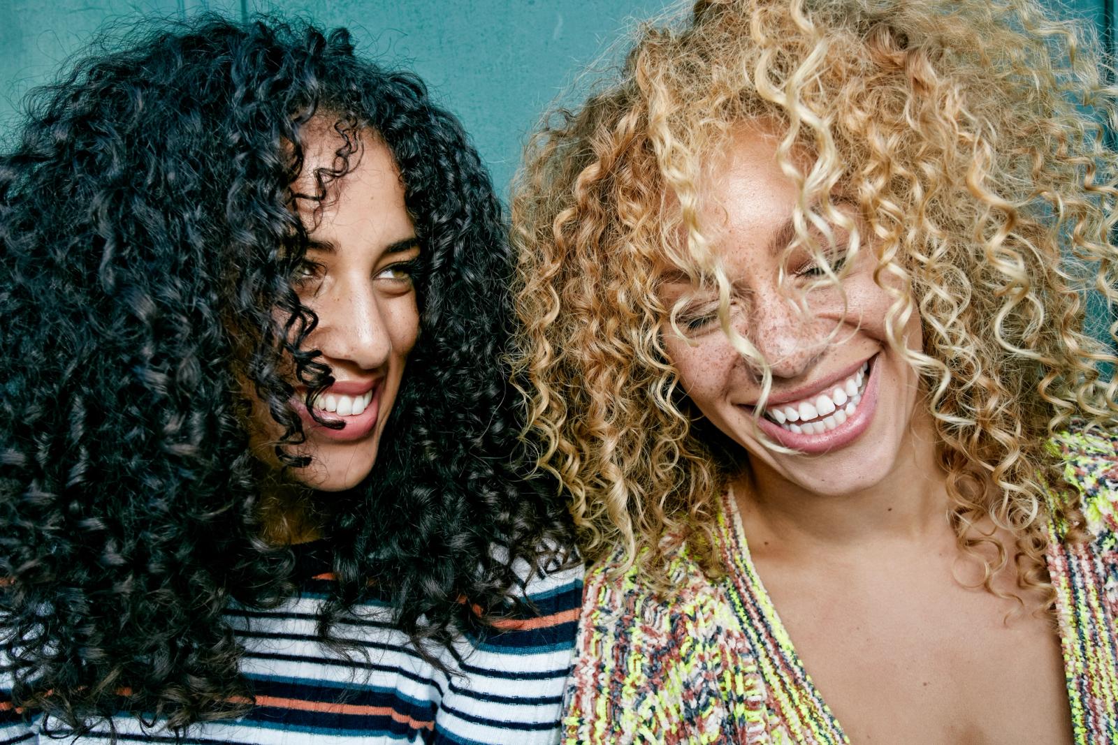 Woman with curly black hair and woman with blonde curly hair laugh standing beside each other