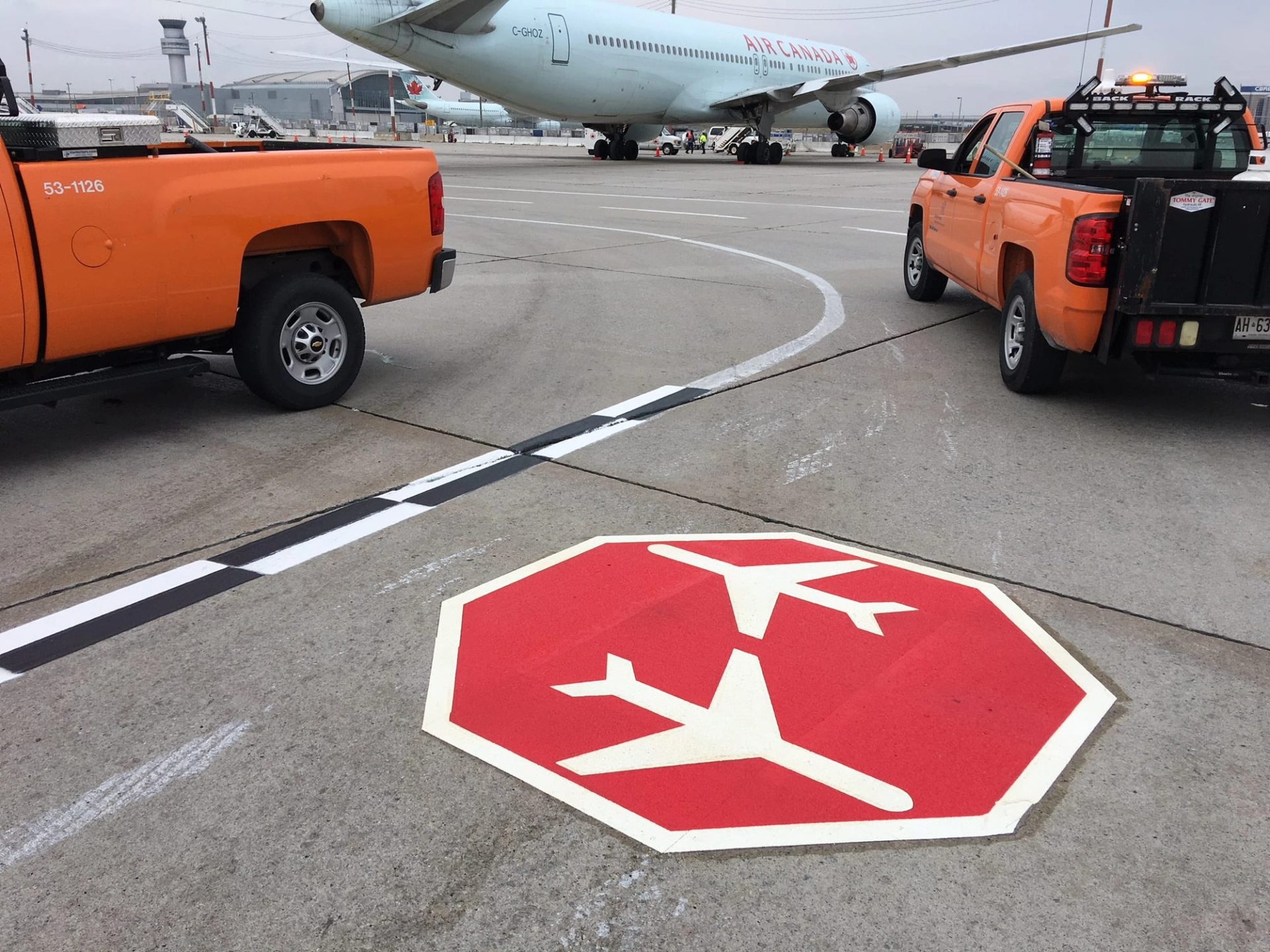 Red octagon marking with white aircraft symbols on an airport apron, orange airside trucks and an Air Canada jet behind