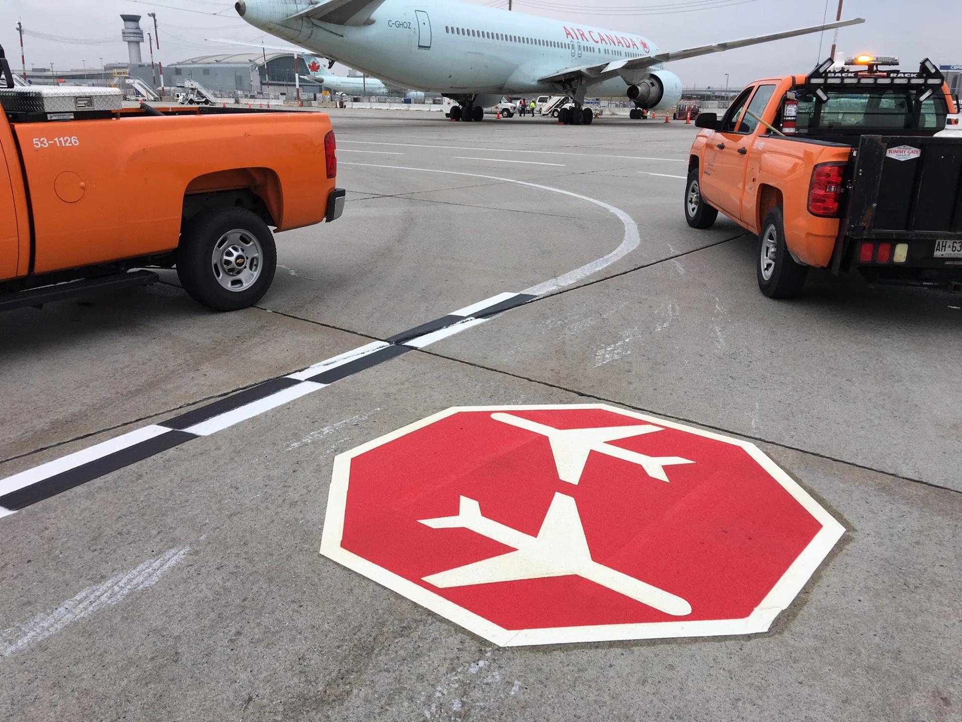 Red octagon marking with white aircraft symbols on an airport apron, orange airside trucks and an Air Canada jet behind