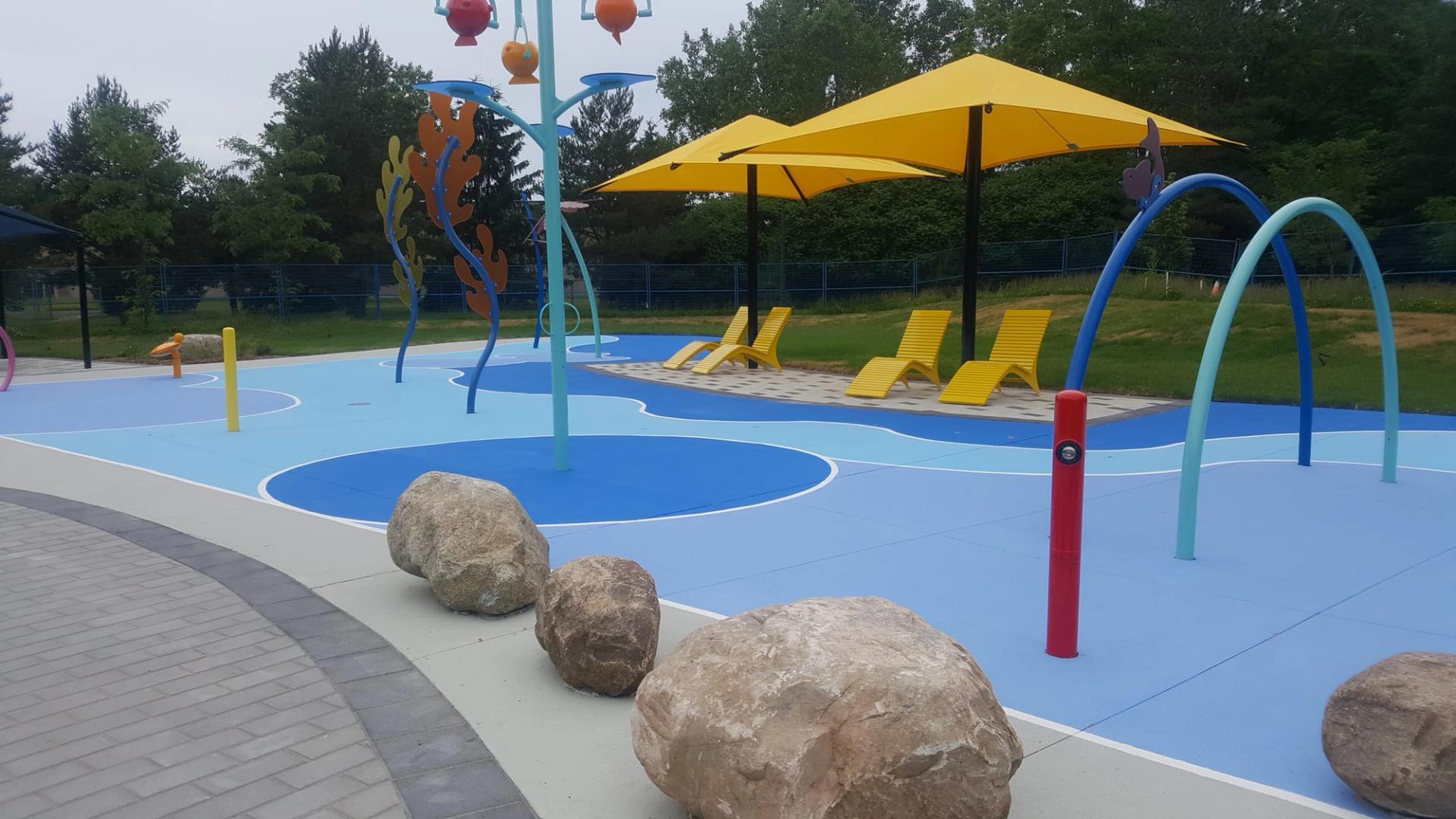 Splash pad surfaced in two shades of blue, with spray arches, boulders and yellow shade umbrellas