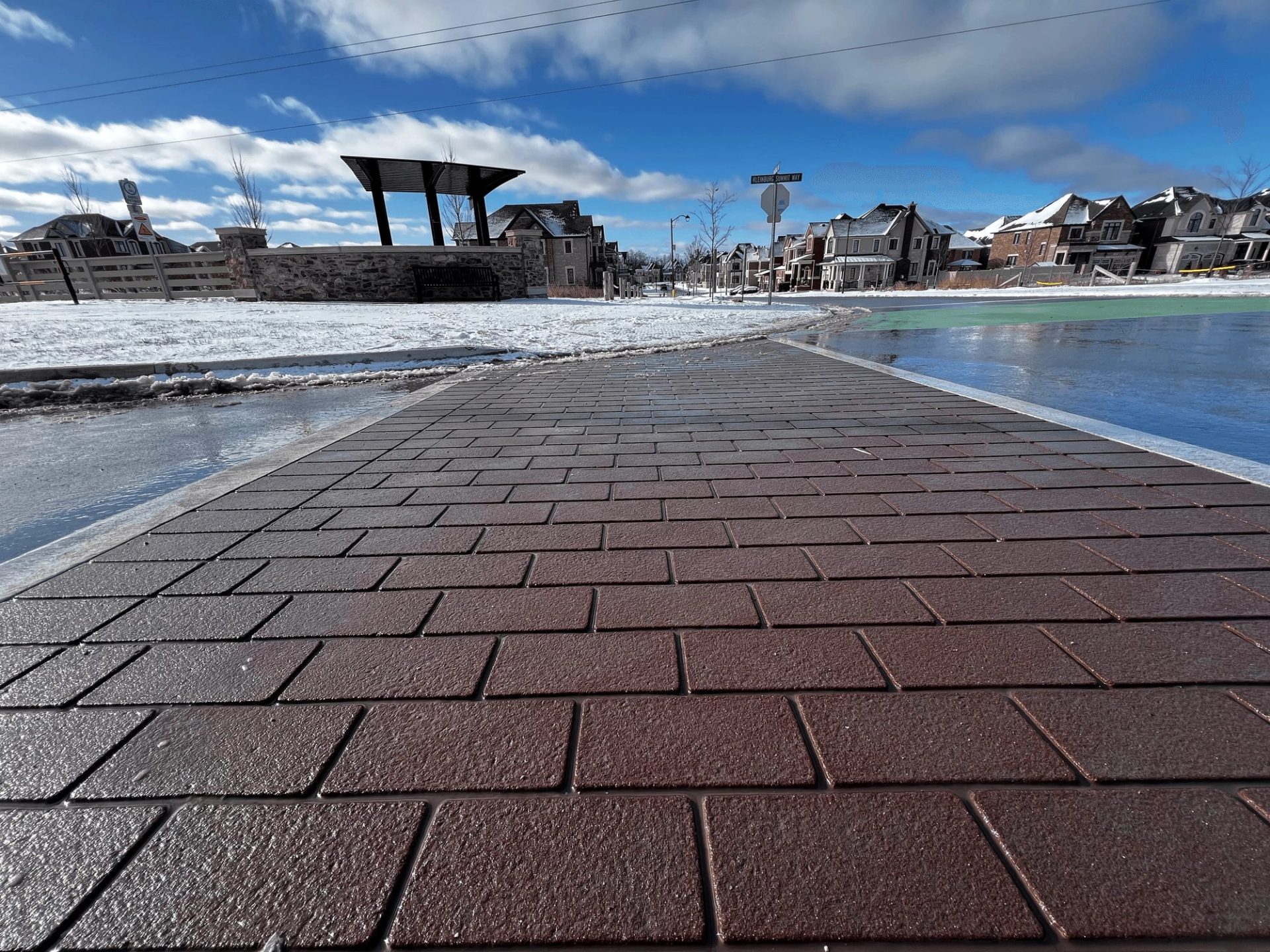 TrafficPatternsXD red brick-pattern walkway after a light snowfall, with a steel shade shelter and new houses behind