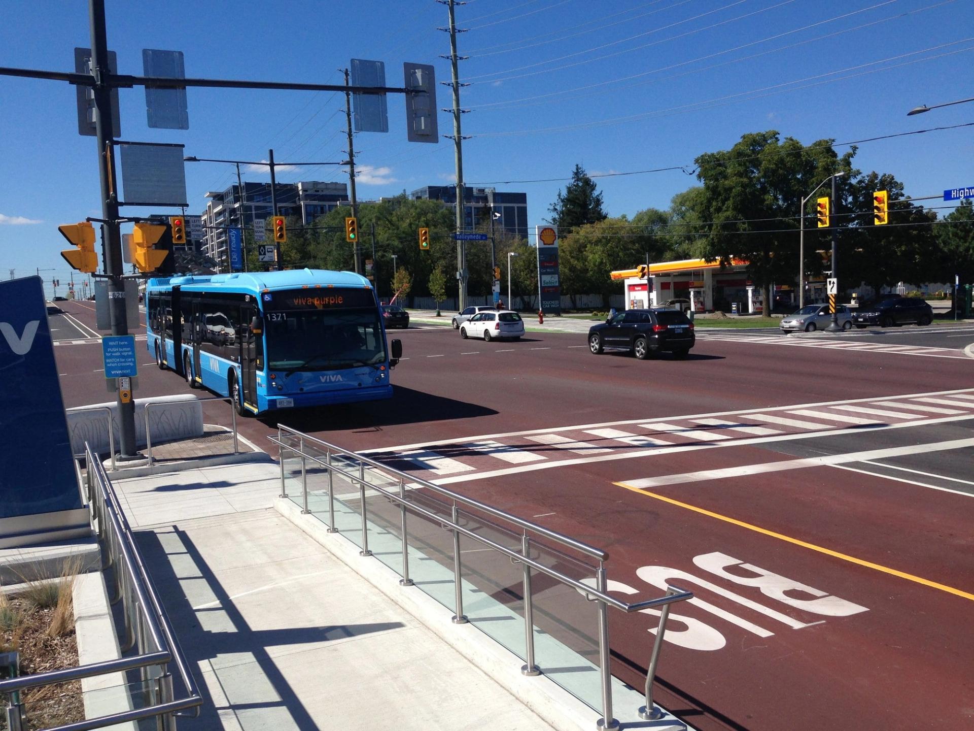 Blue articulated rapid-transit bus on a red bus-only lane at an intersection, beside a station platform