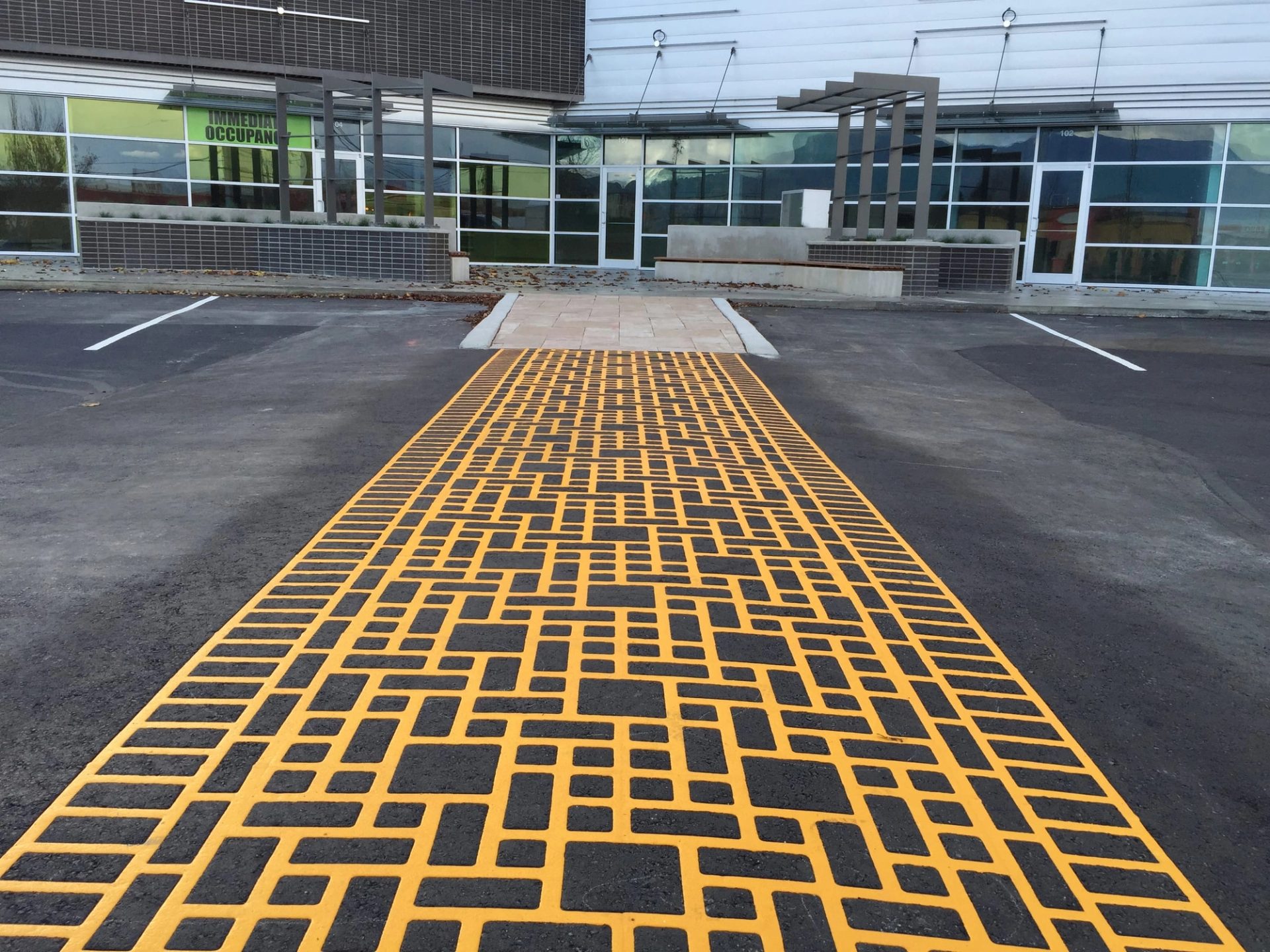 DuraTherm crosswalk inlaid flush in the asphalt, its yellow lines forming a random brick pattern, in front of a glass-fronted building