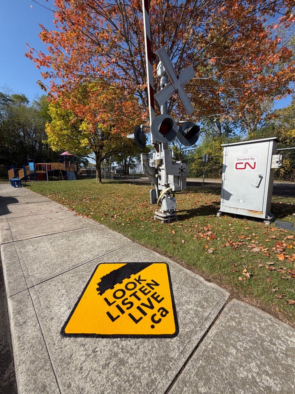 Yellow Look Listen Live DecoMark marking on a concrete sidewalk beside a railway crossing signal and a signal case marked Donated by CN