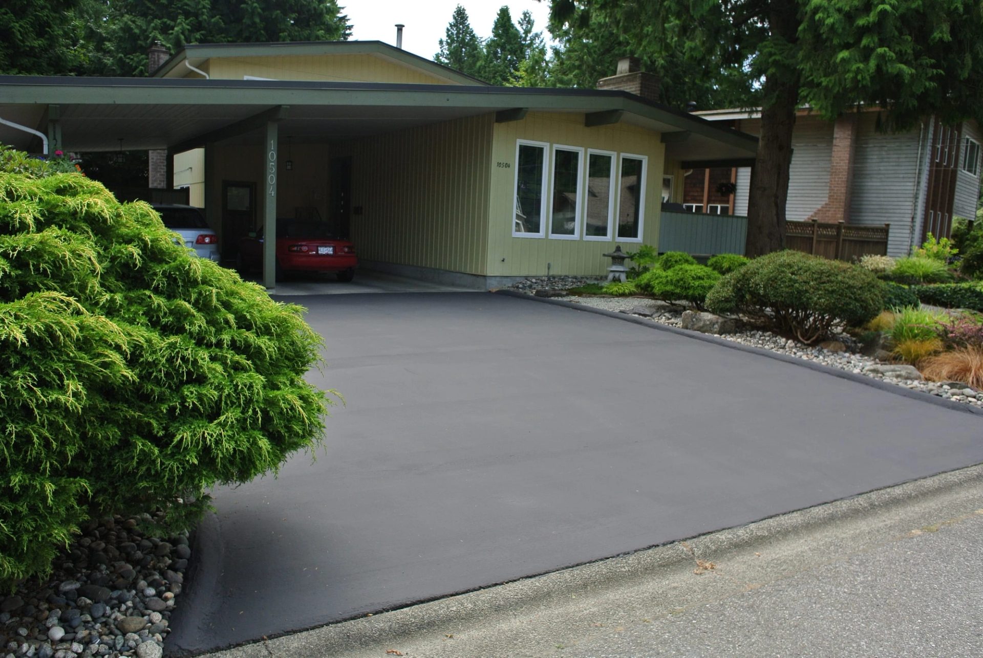 Fresh DuraShield maintenance coating on a residential driveway running from the street to a carport, evergreen shrubs along one side