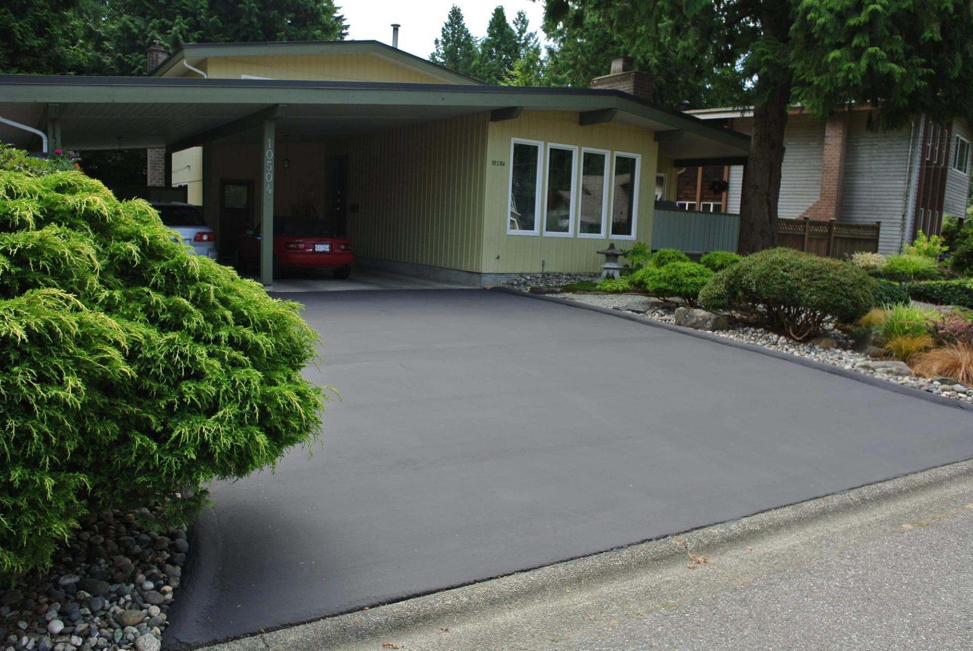 Fresh DuraShield maintenance coating on a residential driveway running from the street to a carport, evergreen shrubs along one side