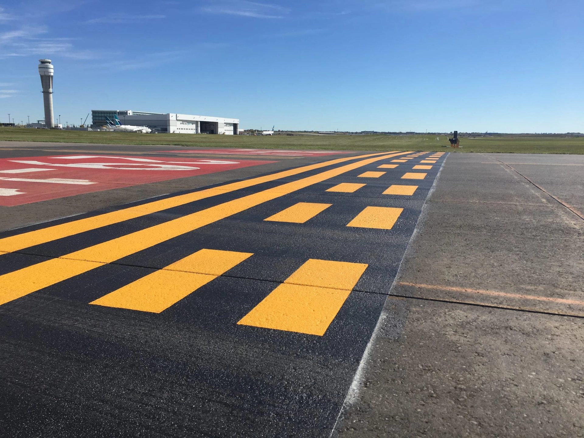 AirMark yellow holding-position lines and a red runway designation marking on an airfield, the control tower and a hangar behind