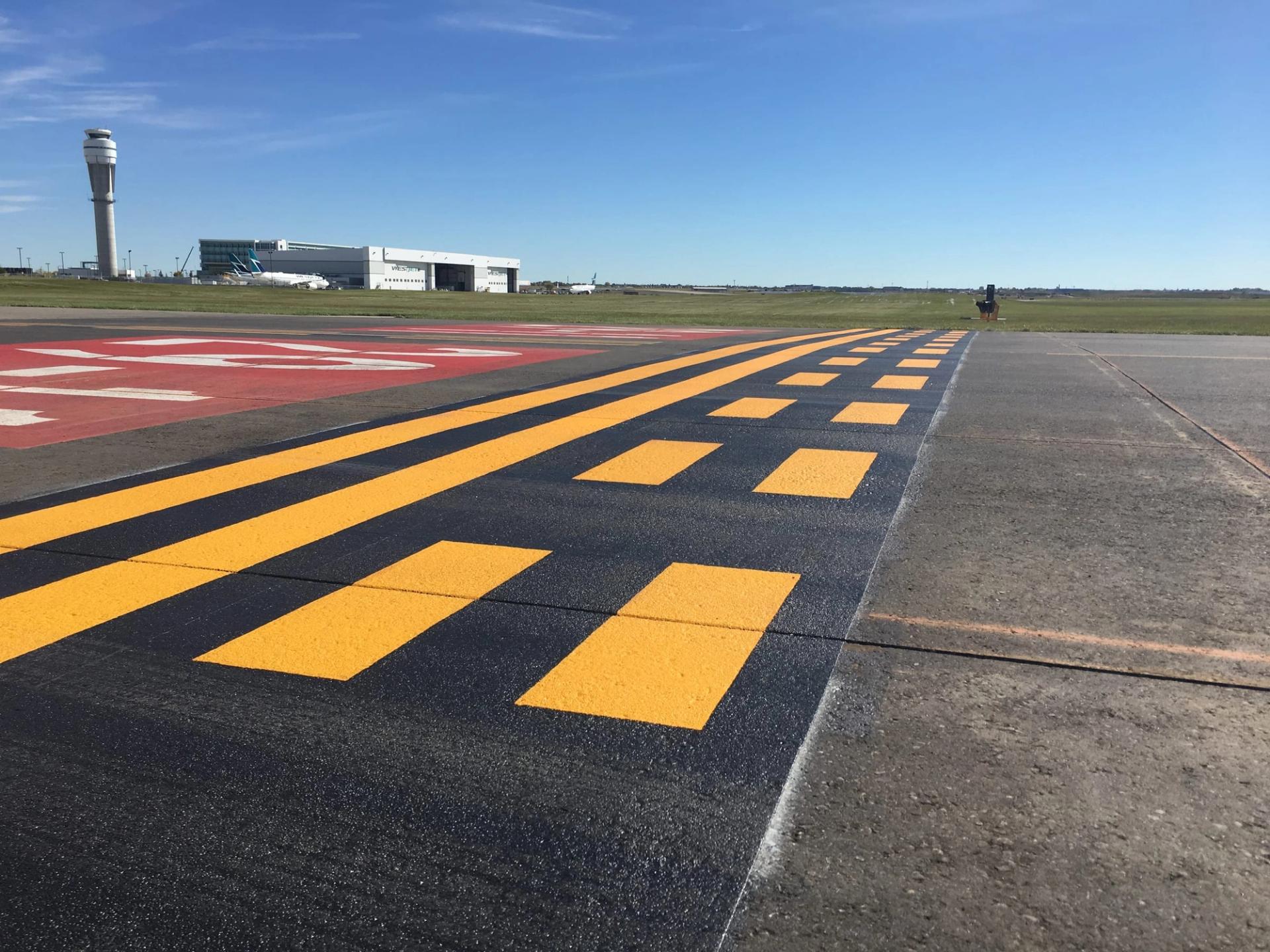 AirMark yellow holding-position lines and a red runway designation marking on an airfield, the control tower and a hangar behind