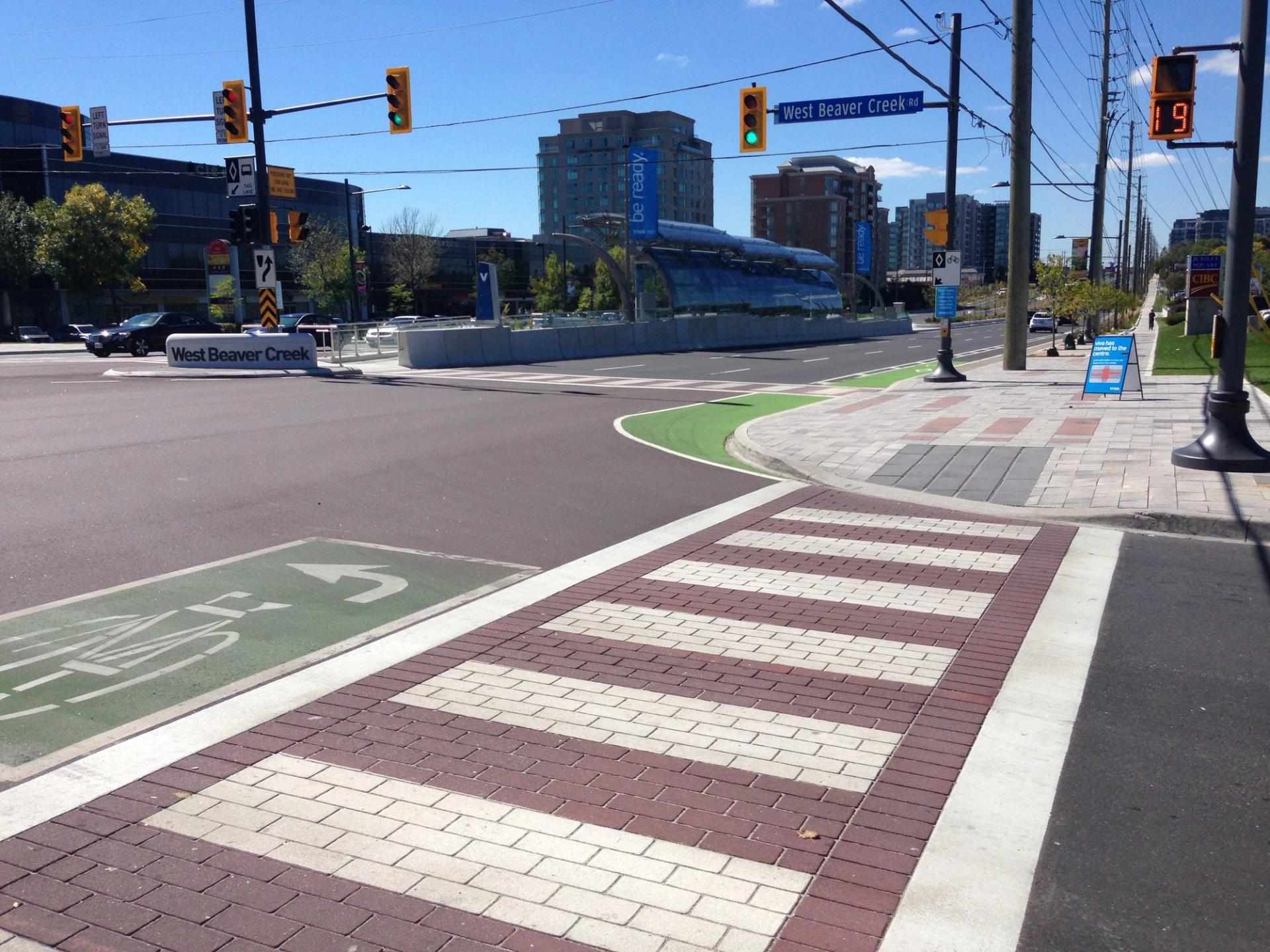 Red and white brick-pattern crosswalk and a green bike crossing at a signalized intersection, office towers behind