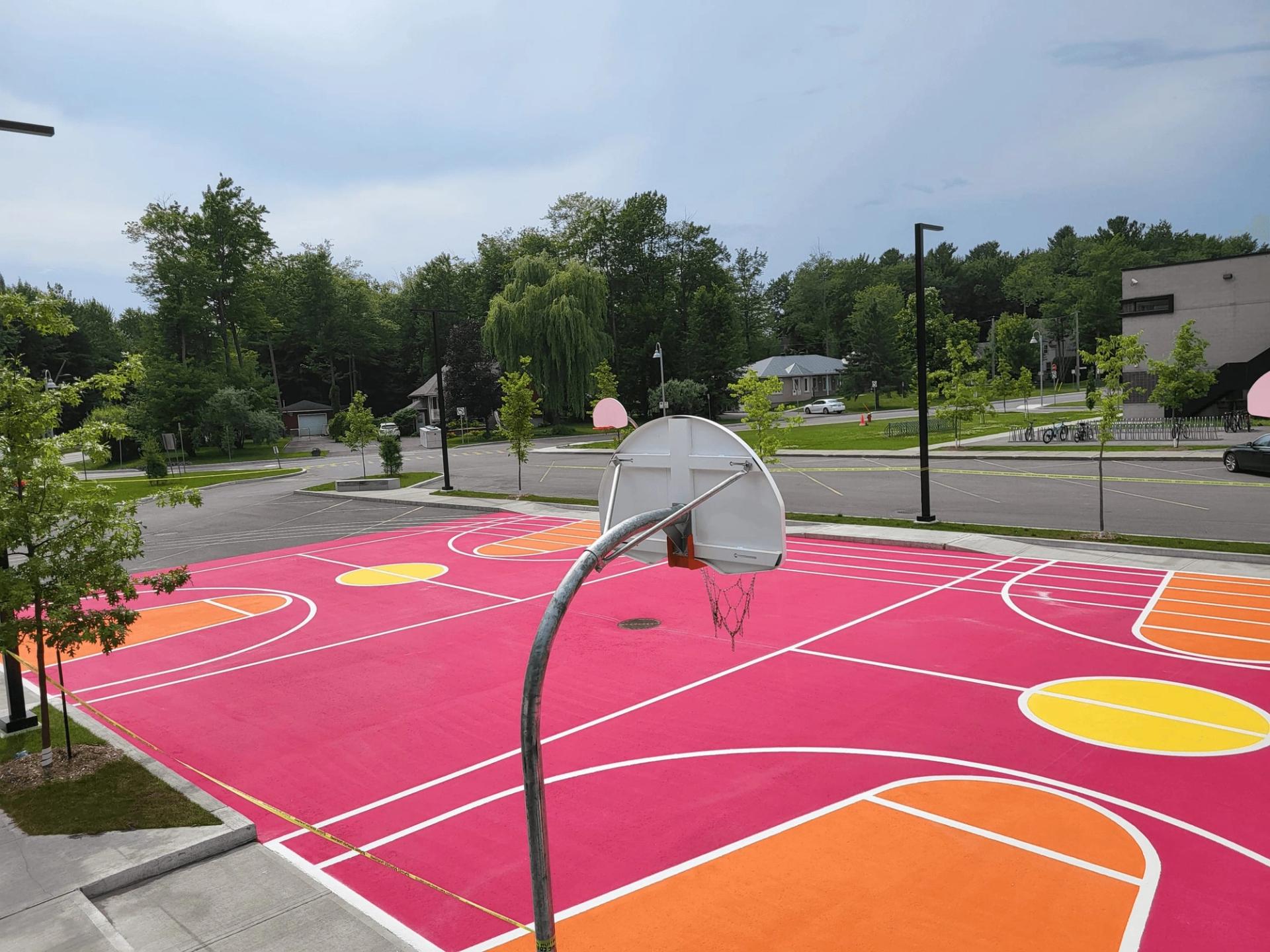 Basketball court coated in pink, orange and yellow StreetBond with white game lines, trees and a street behind
