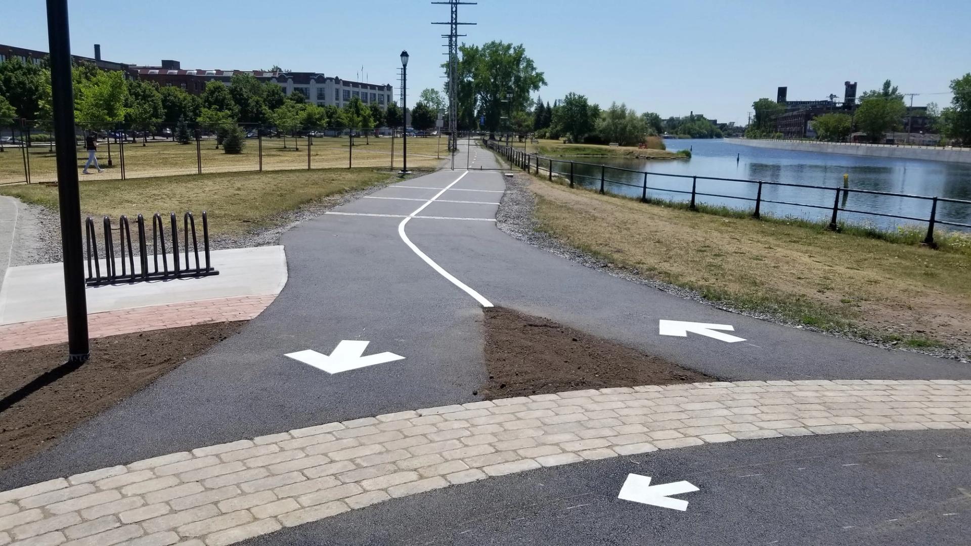 Waterside multi-use path with white direction arrows and a curving centre line, bike racks and a paver edge in the foreground