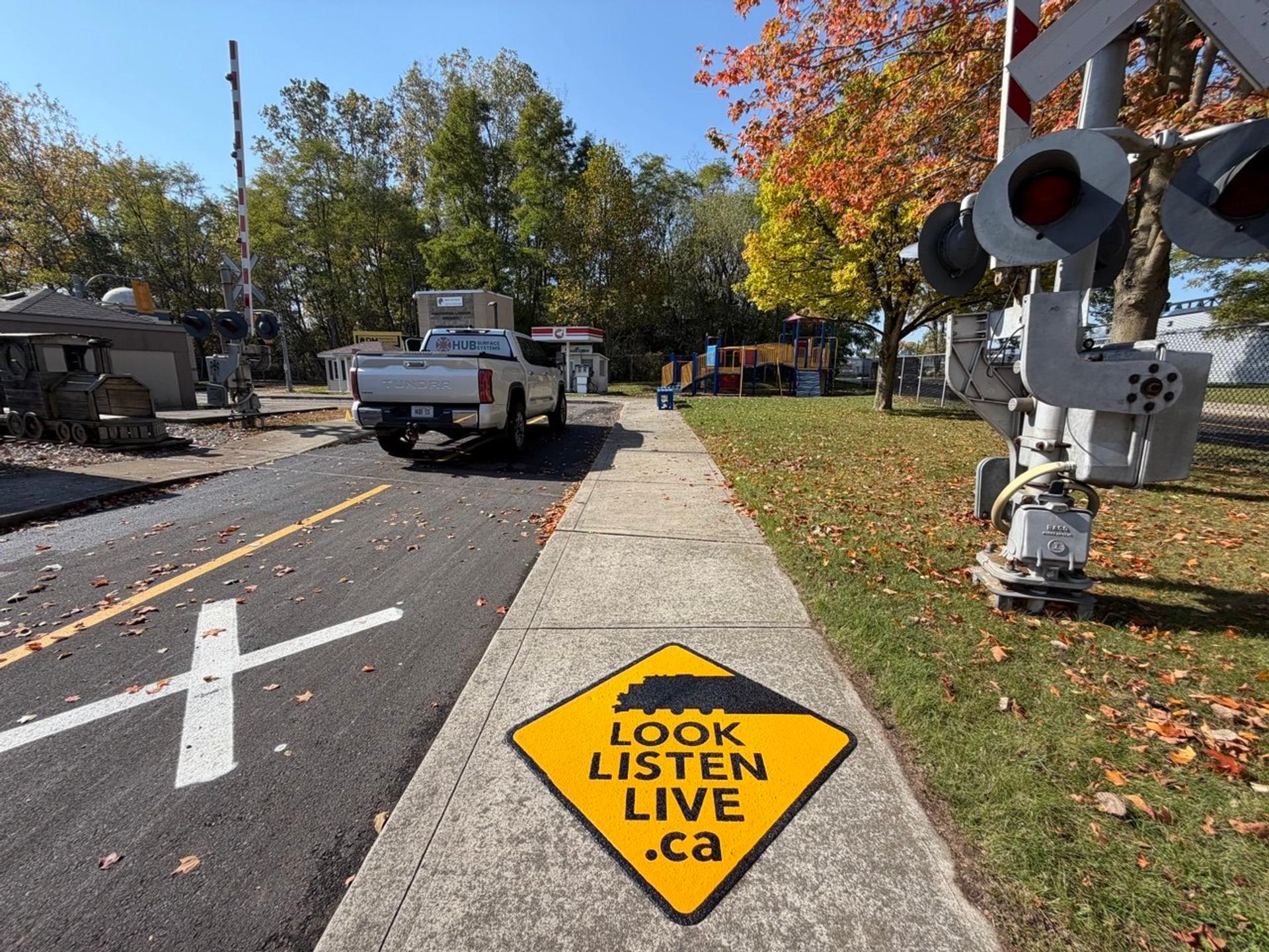 Yellow Look Listen Live rail safety marking on a concrete sidewalk beside a railway crossing signal at the Children's Safety Village of Brant, with a HUB Surface Systems truck behind