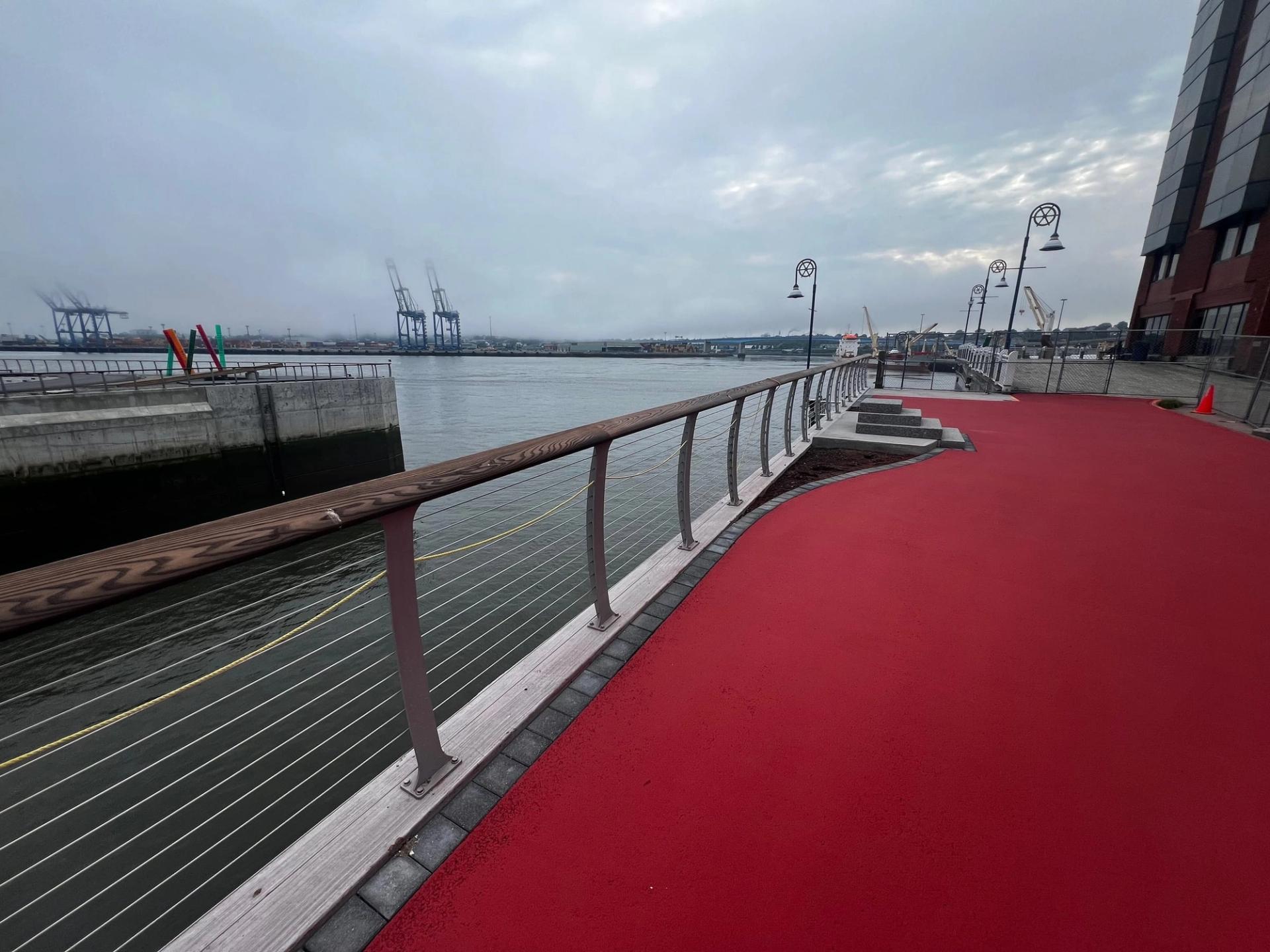 Red StreetBond coating on a harbourfront walkway with a cable railing and lamp posts, port cranes across the water