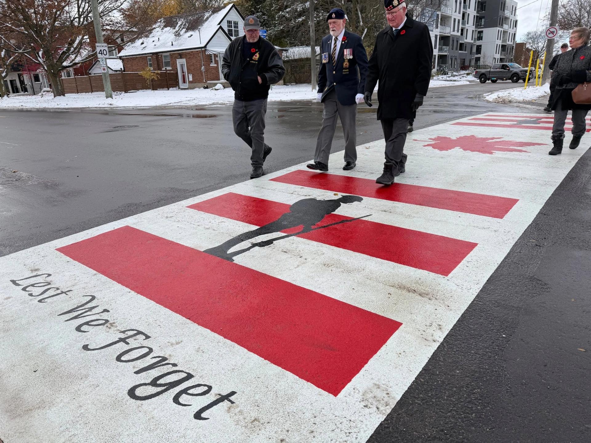 Veterans walk across a TrafficPatterns crosswalk of red bars, a maple leaf and a soldier silhouette above the words Lest We Forget, Kitchener, Ontario