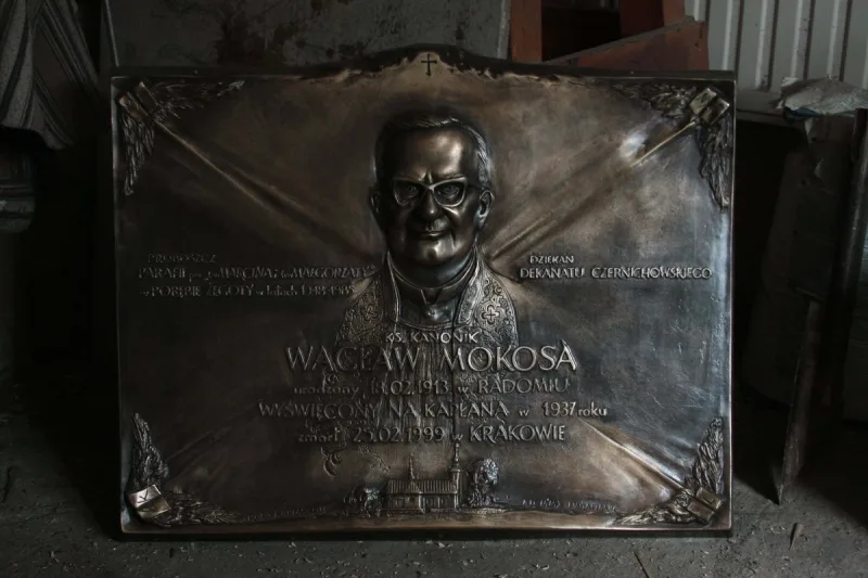 Prostokątna tablica z portretem księdza w okularach i napisami.