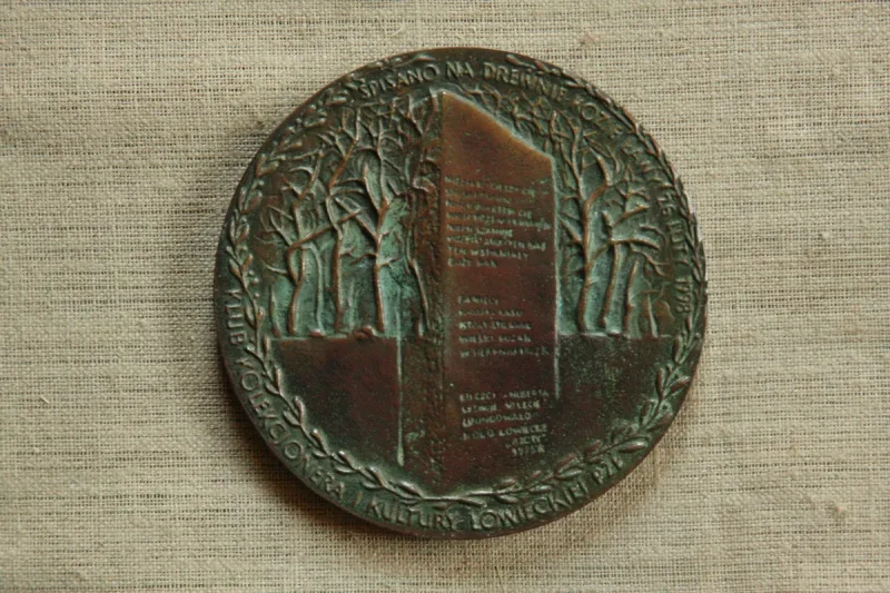 Okrągły medal z pionową tablicą z napisem na tle drzew.