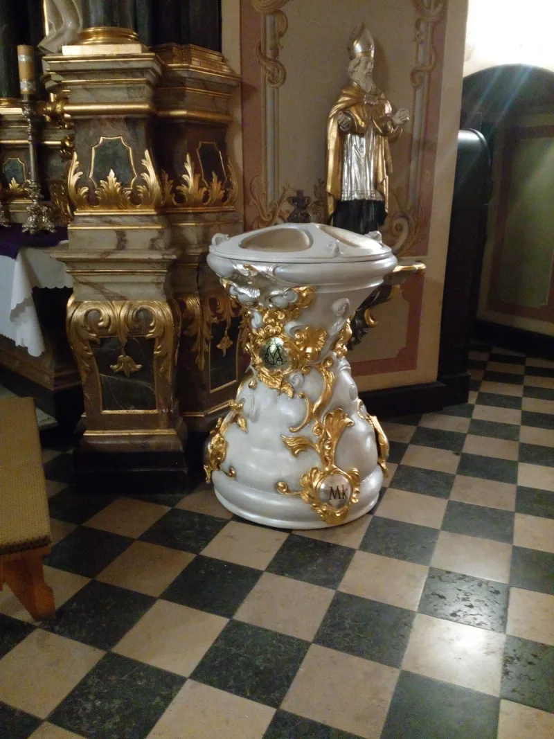 Jasna chrzcielnica z misą i złocistymi dekoracjami, stojąca we wnętrzu kościoła.