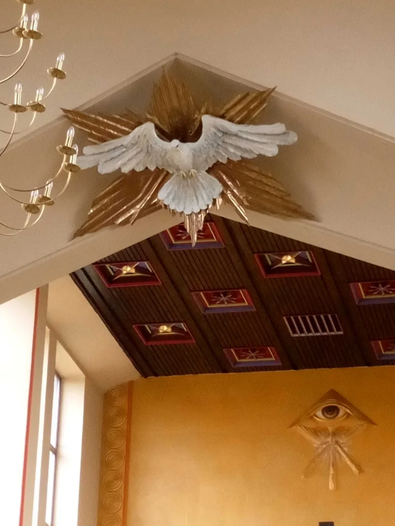 Biały ptak z rozpostartymi skrzydłami na tle złocistych promieni nad wejściem do prezbiterium.