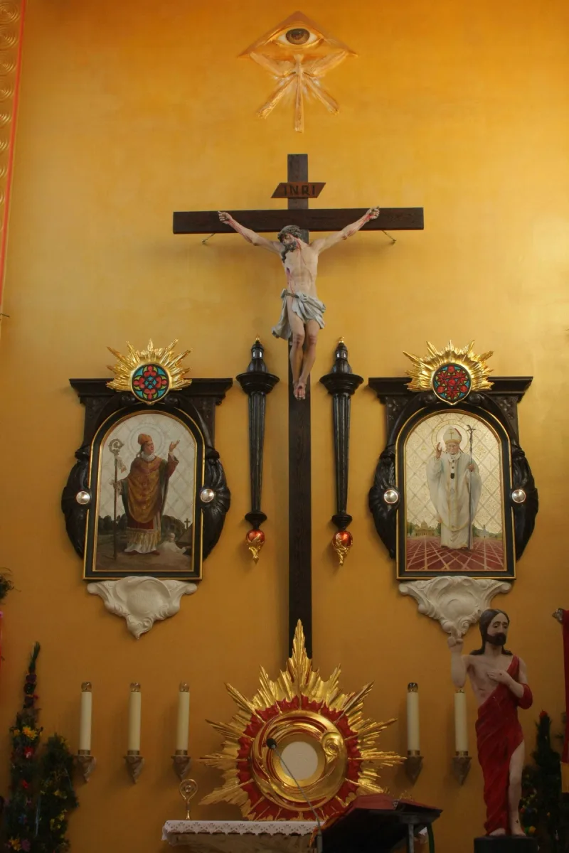 Widok ściany prezbiterium z krucyfiksem, dwoma oprawionymi przedstawieniami i tabernakulum.