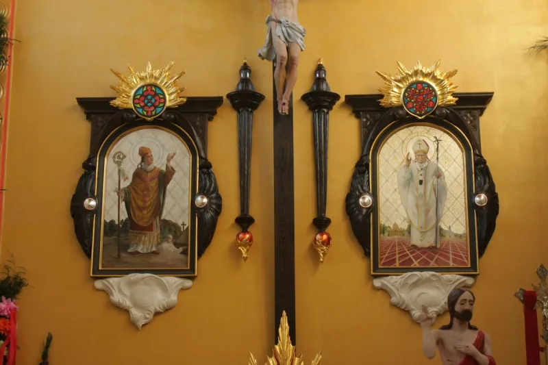 Ściana prezbiterium z krucyfiksem i dwoma przedstawieniami w ozdobnych ramach.