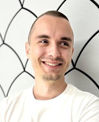 Smiling man in a white Adidas T-shirt stands with folded arms against a light wall with a black scallop pattern.