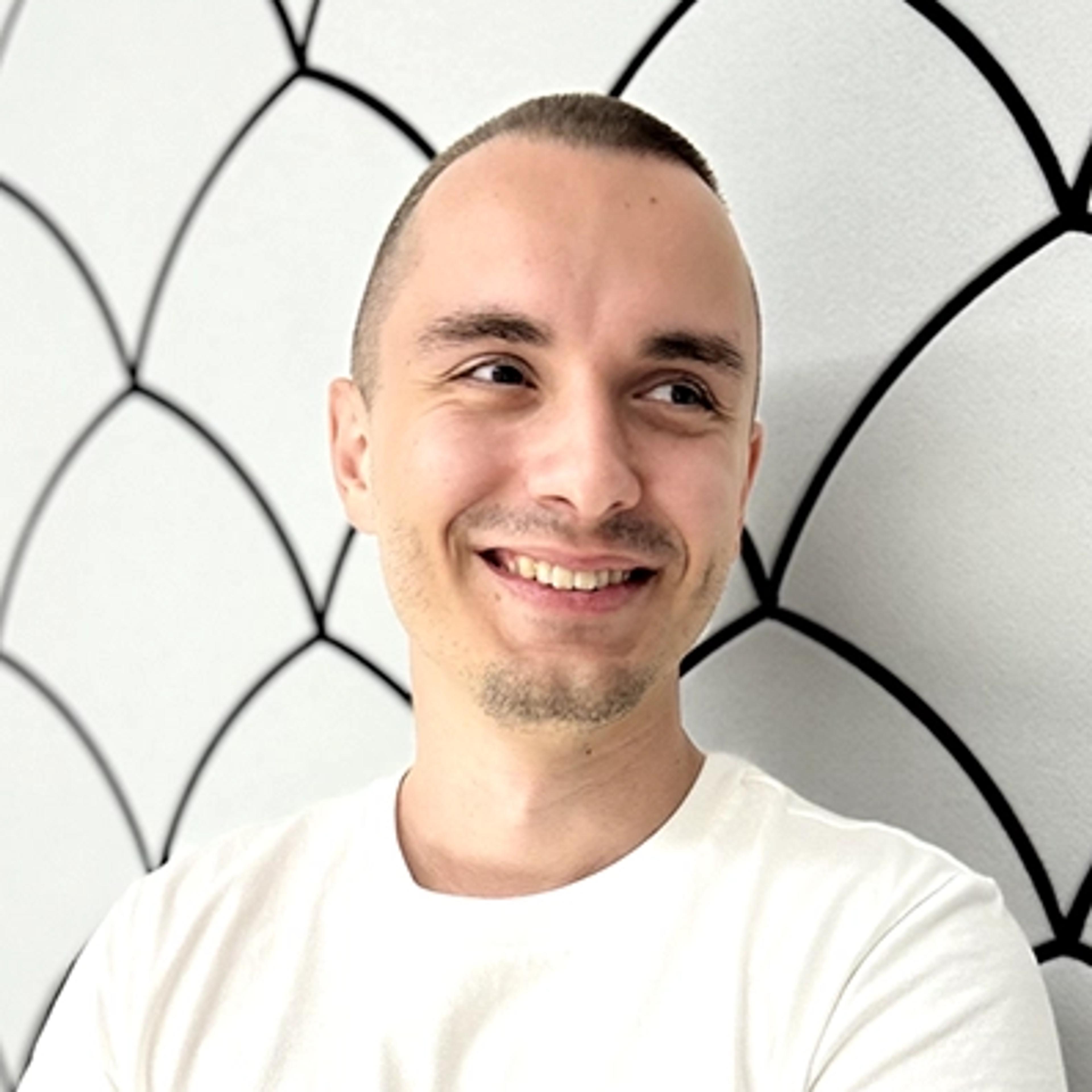 Smiling man in a white Adidas T-shirt stands with folded arms against a light wall with a black scallop pattern.