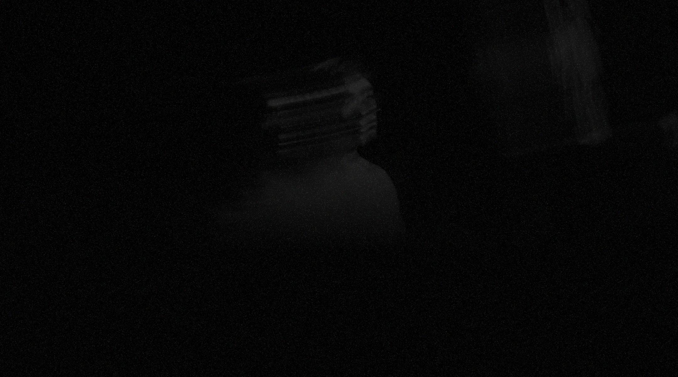 Dark, grainy scene with a faint, blurred figure in a white shirt emerging from the shadows