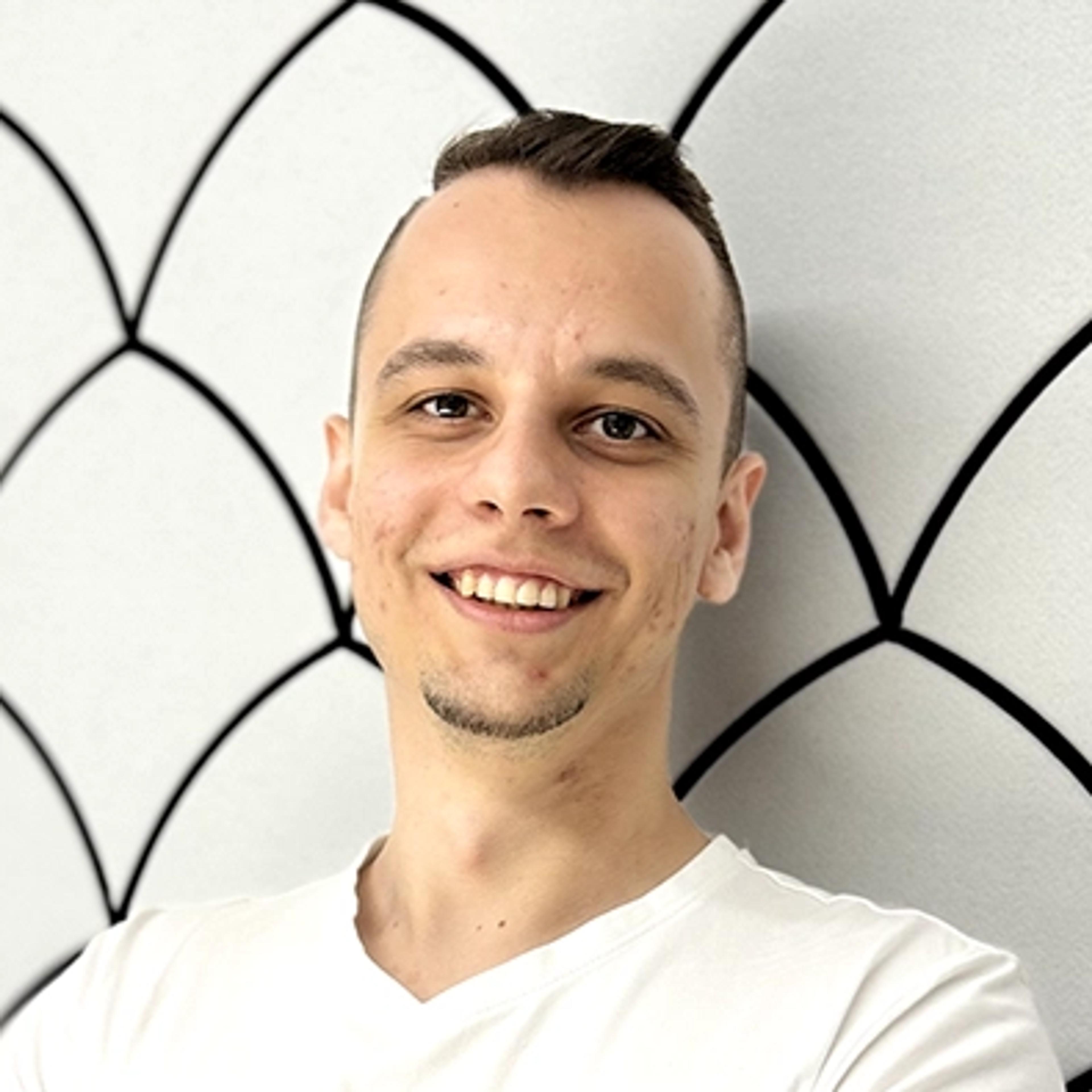 Smiling man in a white T-shirt with arms crossed, standing against a white wall with black geometric patterns.