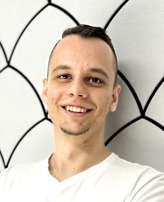 Smiling man in a white T-shirt with arms crossed, standing against a white wall with black geometric patterns.