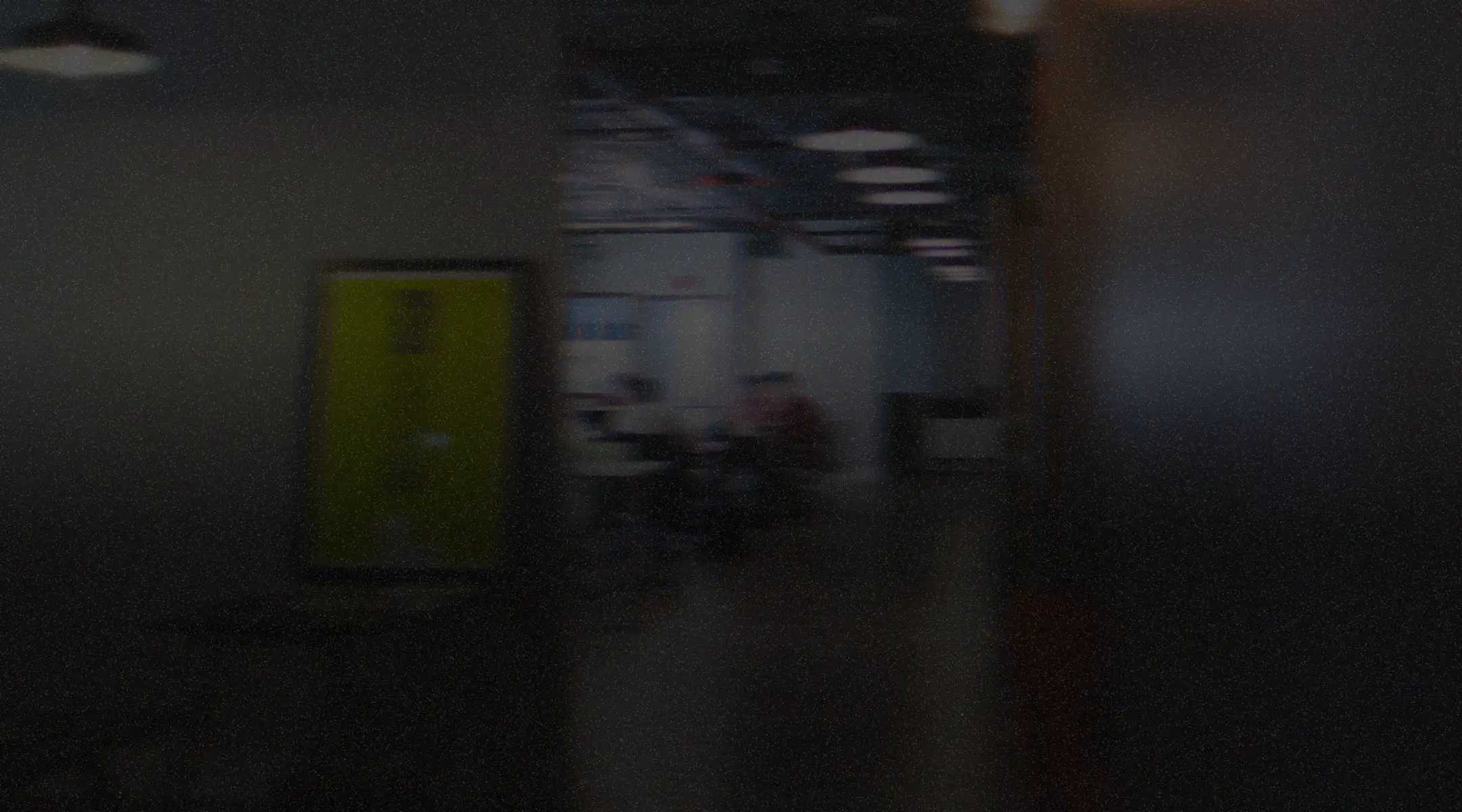 Dimly lit office hallway with a yellow door and people working at desks in the background.