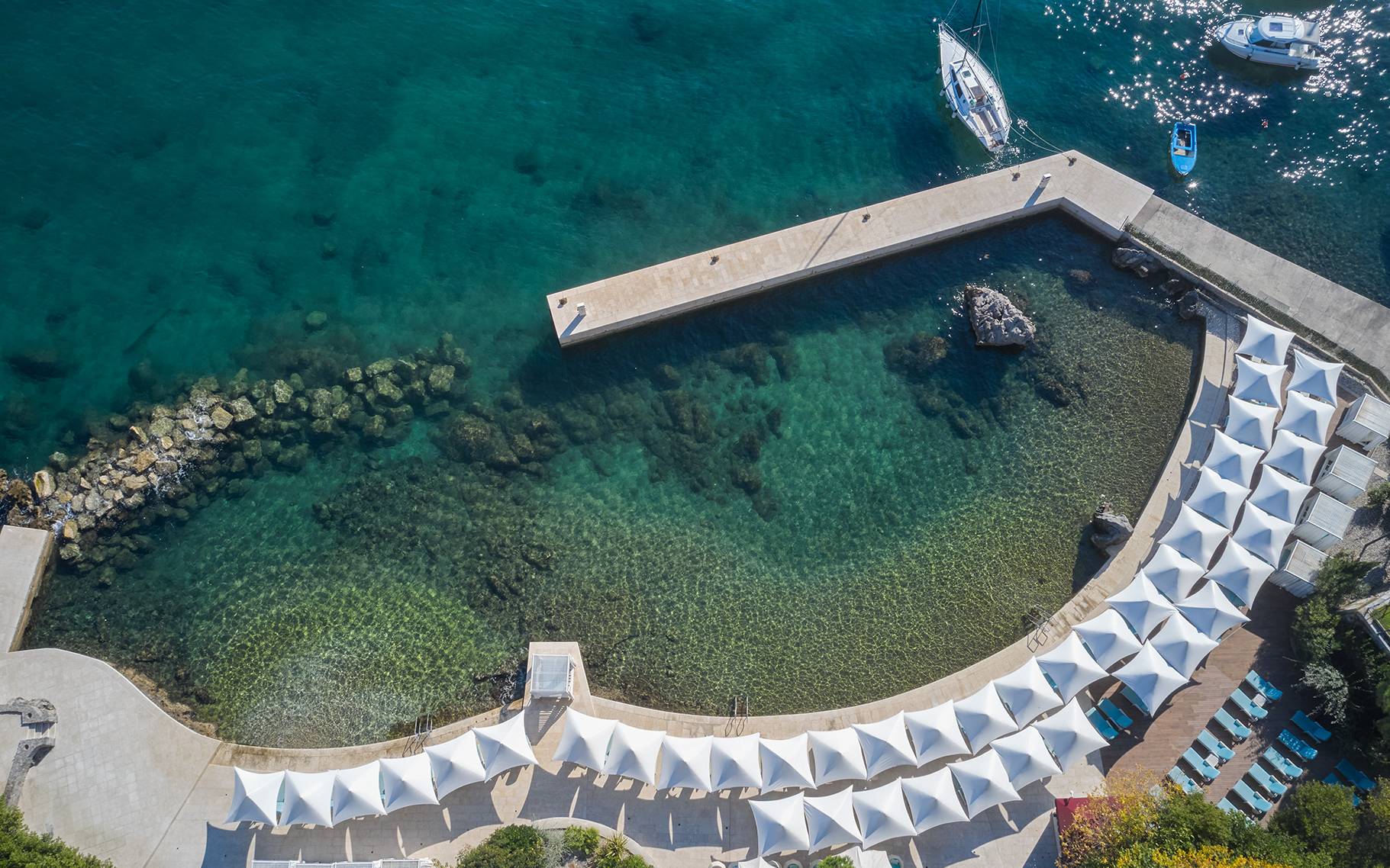 Royal Beach iz zraka: bijela sjenila u obliku jedra i plave ležaljke uz zakrivljenu kupališnu platformu nad tirkiznim morem