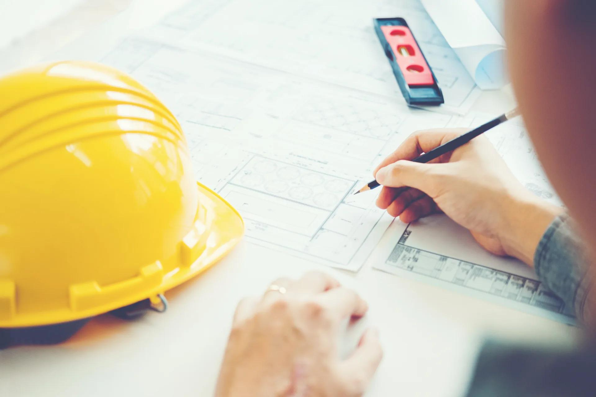 Engineer marking up a set of construction drawings by hand at a review table, with a hard hat and level resting on the plans