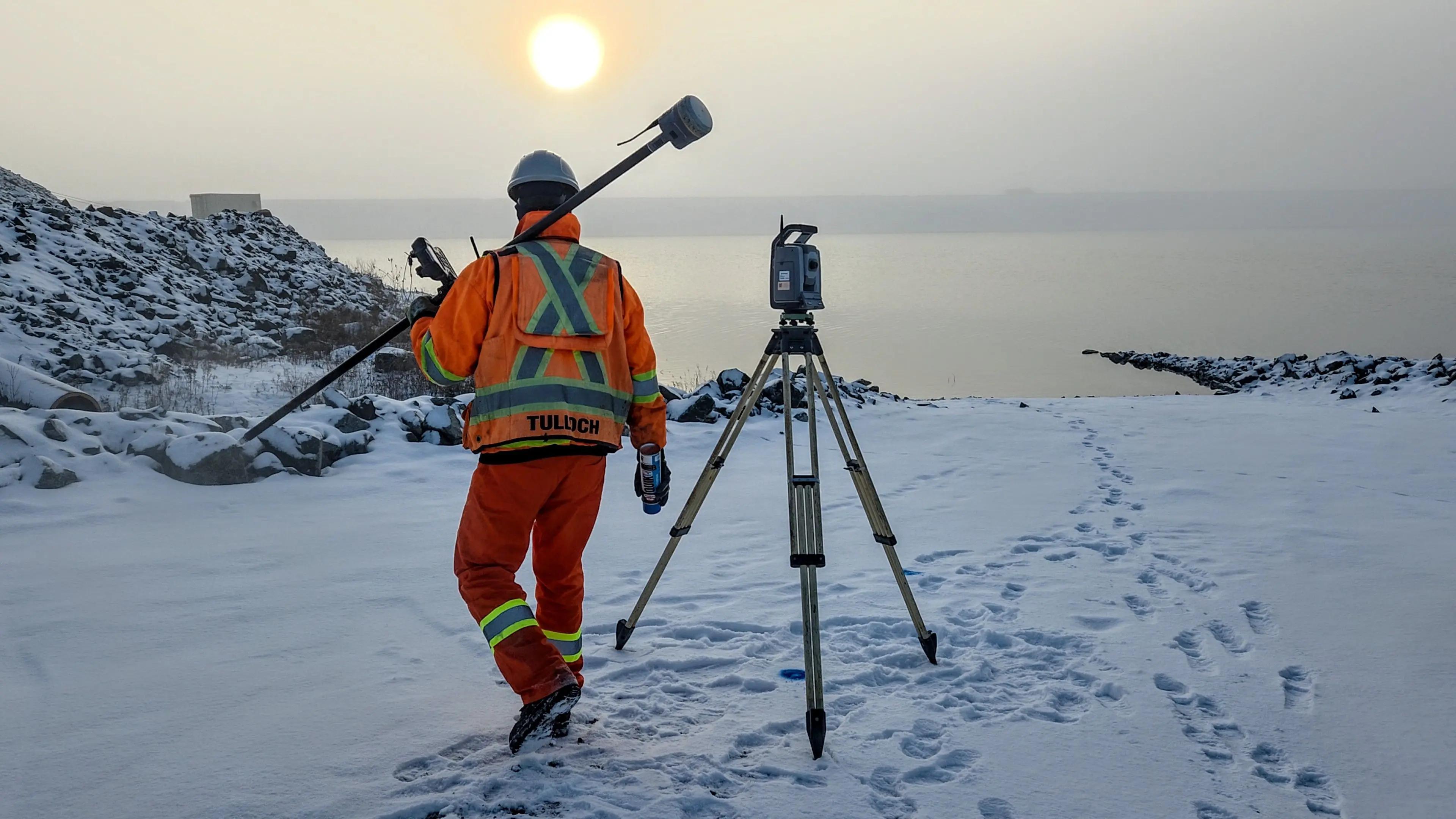 TULLOCH surveyor carrying field equipment toward a total station on a snow-covered site.