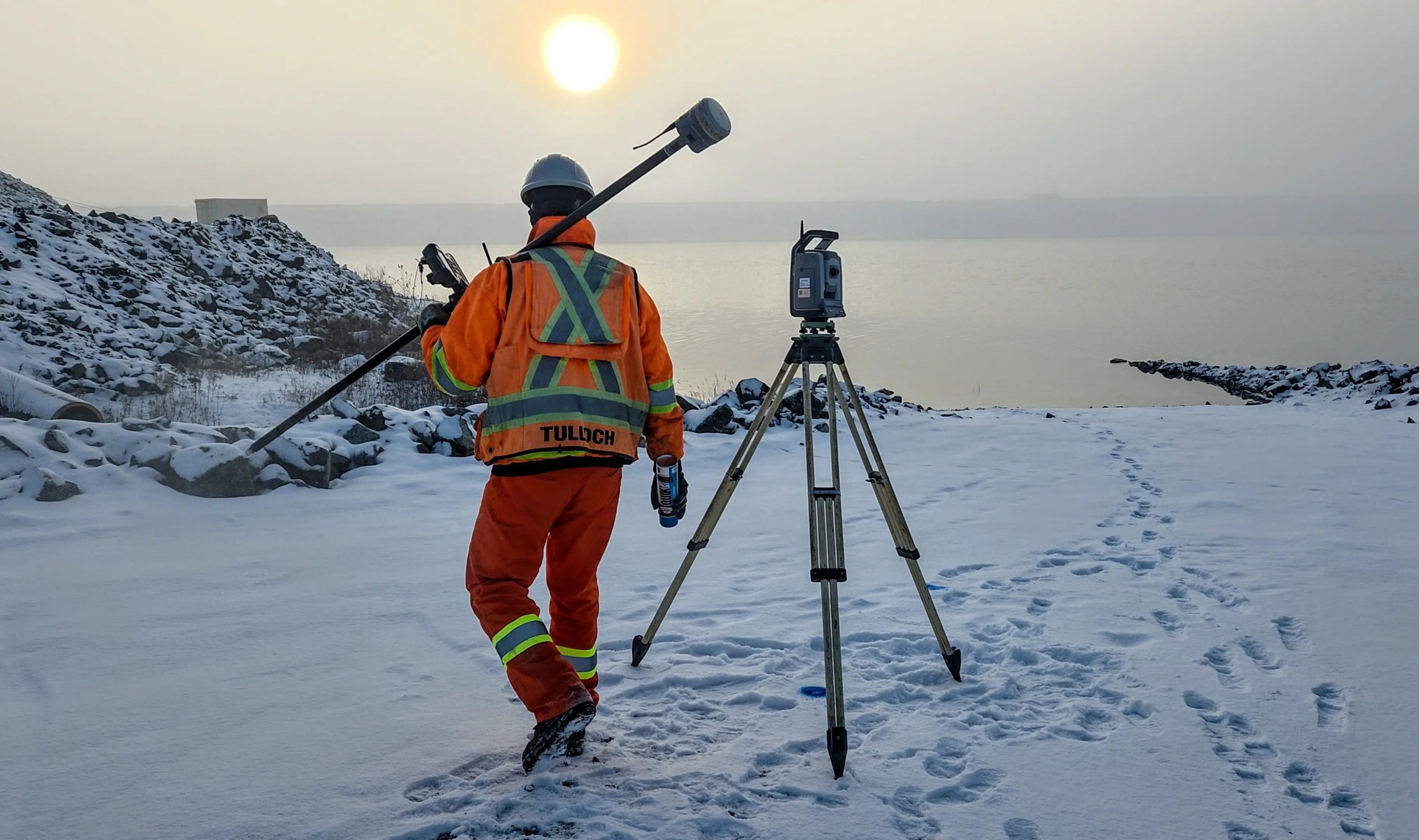 TULLOCH surveyor carrying field equipment toward a total station on a snow-covered site.