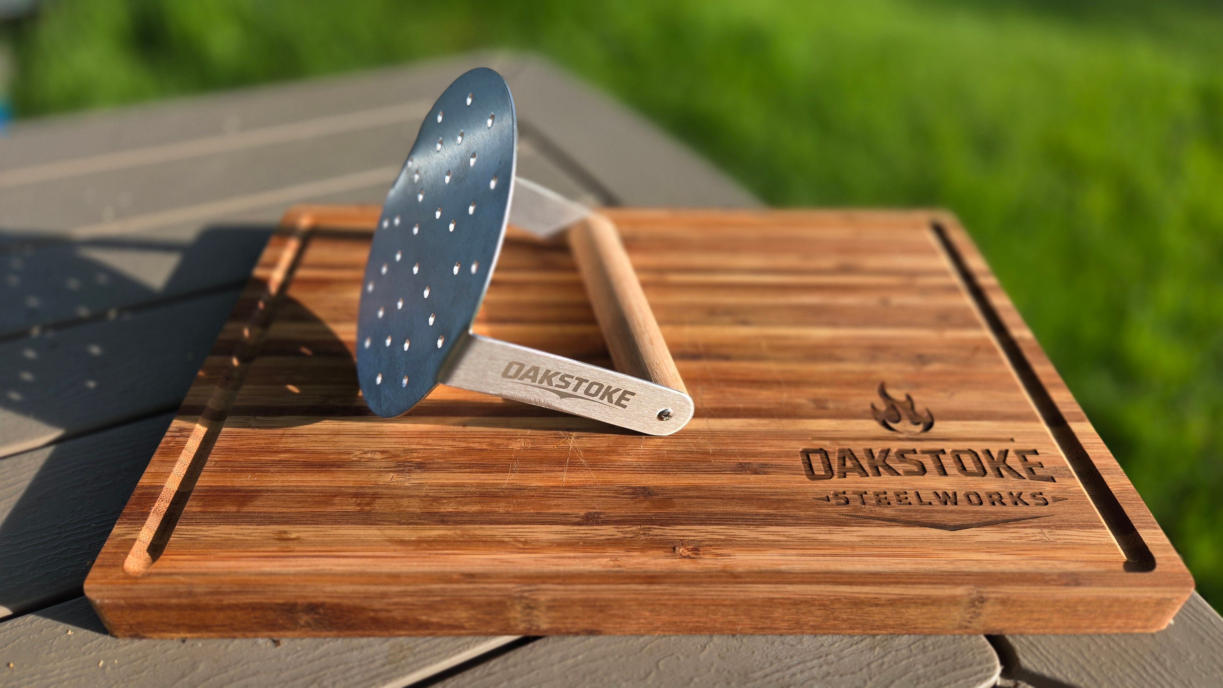 Alt text: OakStoke Steelworks stainless steel smash burger press with a perforated round head and wood handle, standing upright on an engraved bamboo cutting board outdoors.