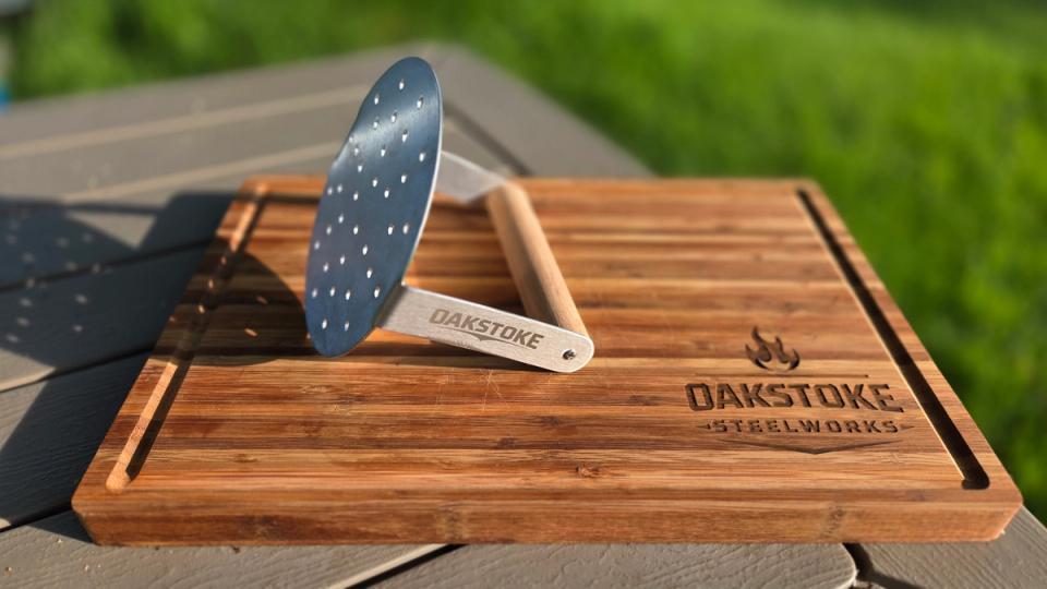OakStoke Steelworks stainless steel smash burger press with a perforated round head and wood handle, standing upright on an engraved bamboo cutting board outdoors.