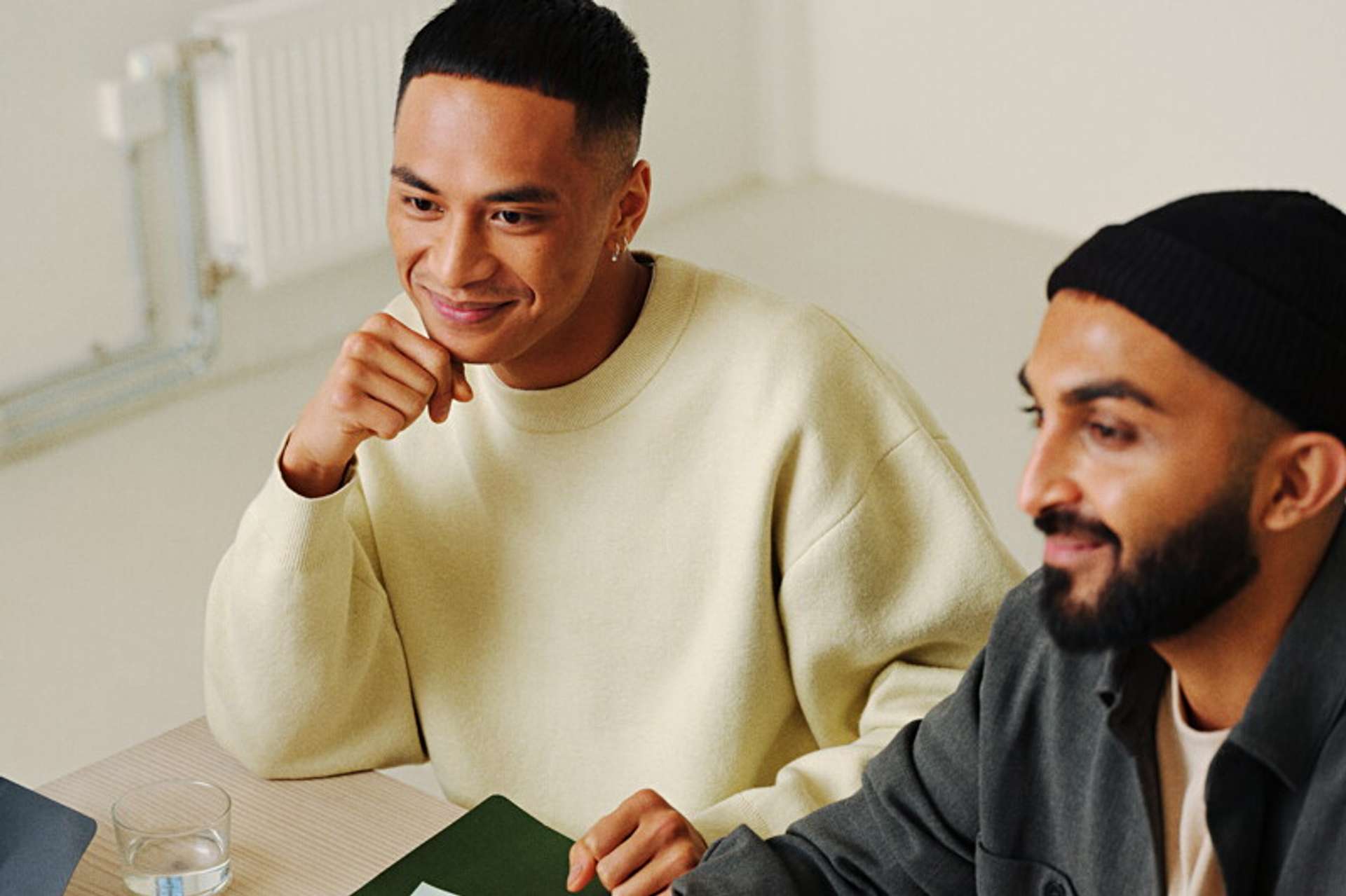 Two men smiling around a table, one wearing a yellow jumper and supporting his chin with his hand, the other wearing a black beanie
