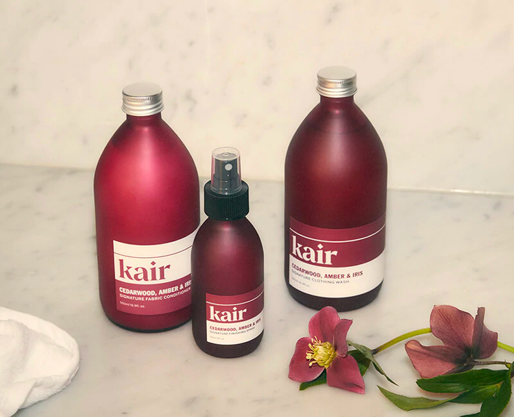 Three red glass bottles with rectangle stickers printed with the Kair logo and information on the laundry detergents. The bottles are on a marble counter top with a flower and towel laying nearby.