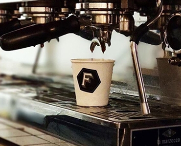 Compostable coffee cup printed with the Foundation logo. The coffee cup is being filled with coffee in an espresso machine.