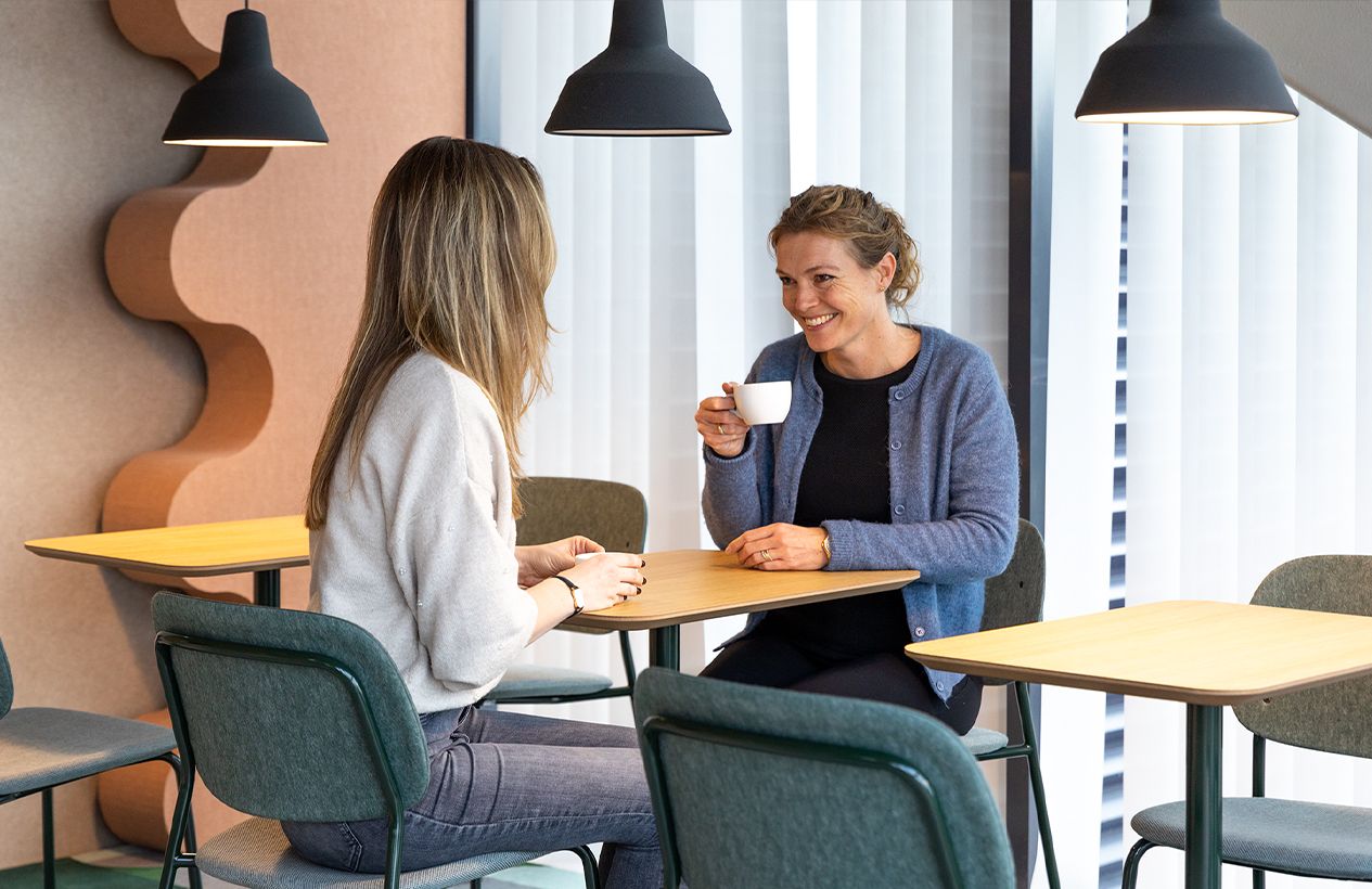Two women in conversation at a small oak table, seated on Hale Stack Chairs in dark green PET felt in a bright office breakout area