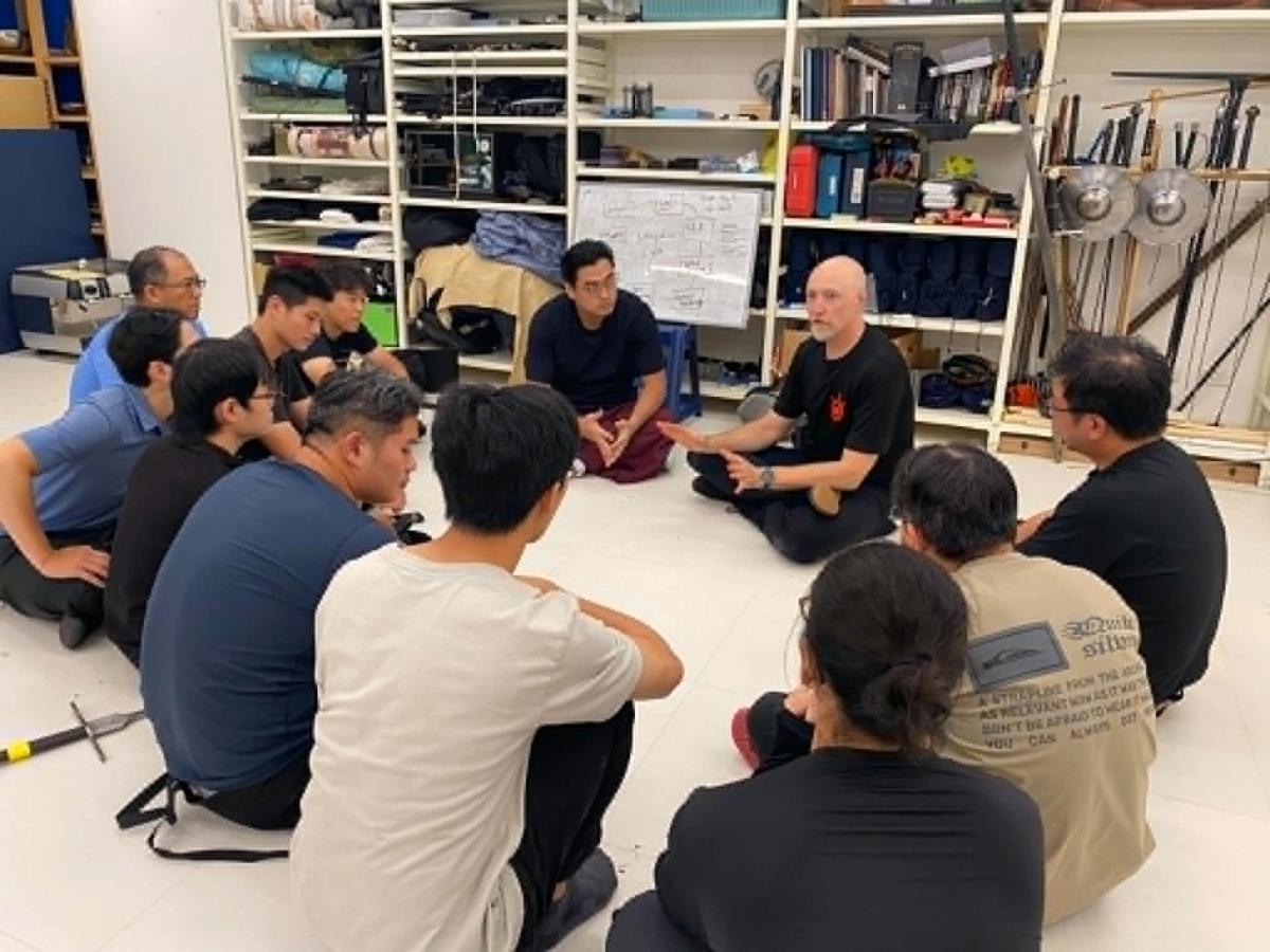 Intensive participants sitting on the floor in a discussion circle, with racks of training swords and gear behind them