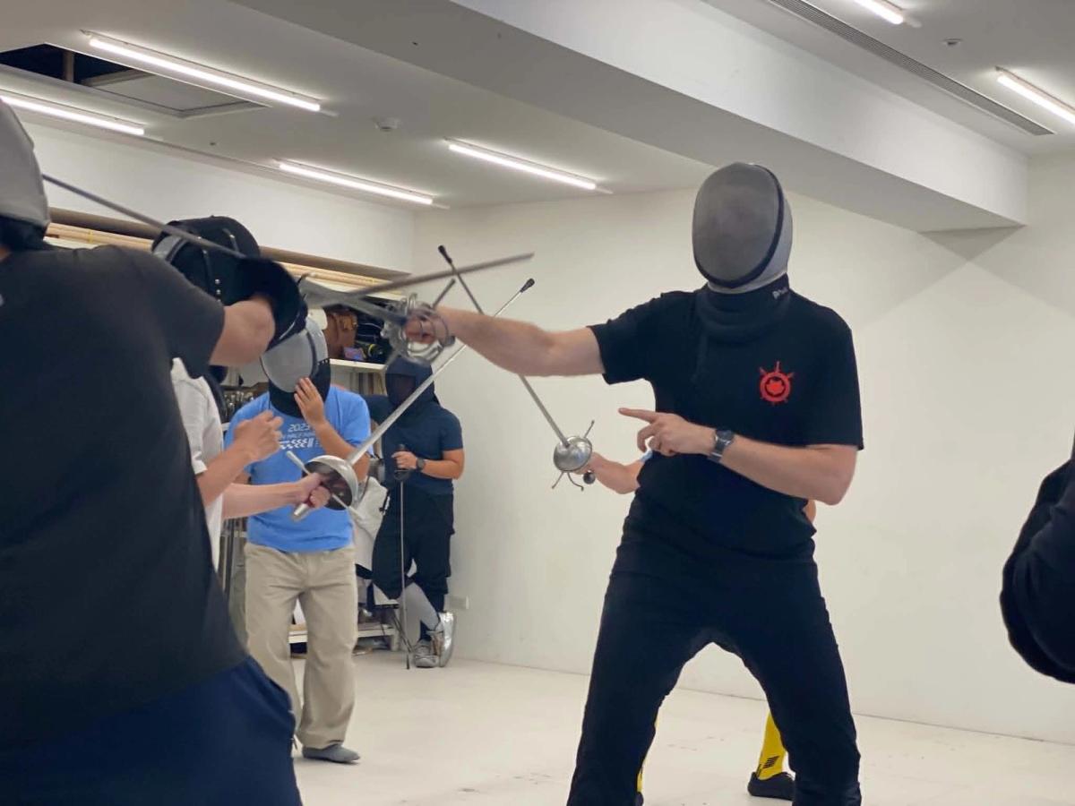 Devon Boorman, in a fencing mask, demonstrates a rapier bind with a student while other participants look on