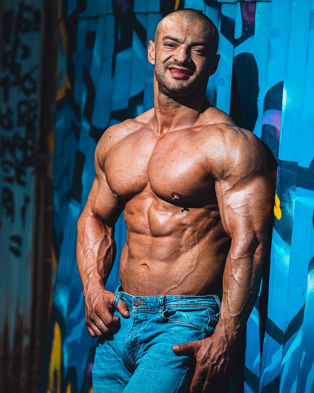 Urban fitness portret muškarca sa naglašenom mišićnom definicijom, snimljen uz zid sa grafitima, sa snažnim kontrastnim osvetljenjem i fokusom na snagu, teksturu i karakter.