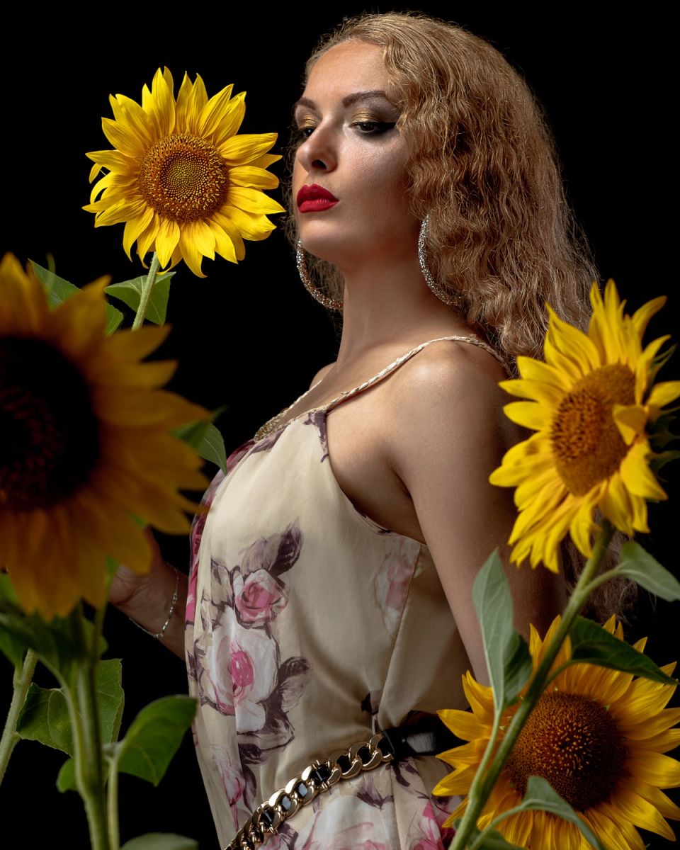 Portret žene u elegantnoj haljini okružene suncokretima, na tamnoj pozadini