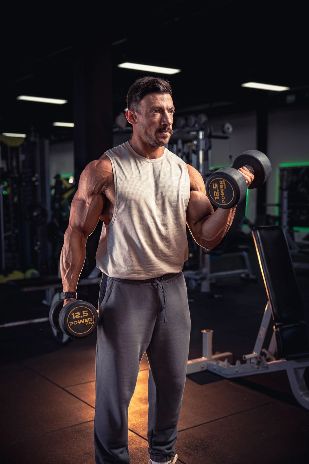 Fitness portret muškarca tokom treninga sa bučicama u teretani, sa naglašenom muskulaturom, dramatičnim osvetljenjem i fokusom na snagu i formu.
